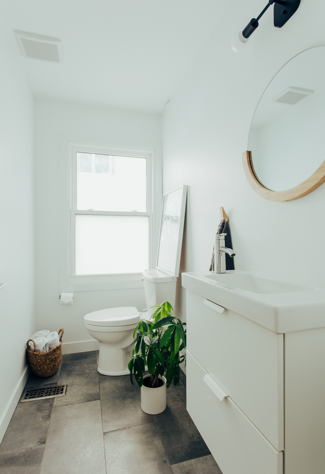 Get small bathroom inspiration from this bright powder room featuring a white vanity, round mirror with a wood accent, grey floor tiles, and a frosted window.