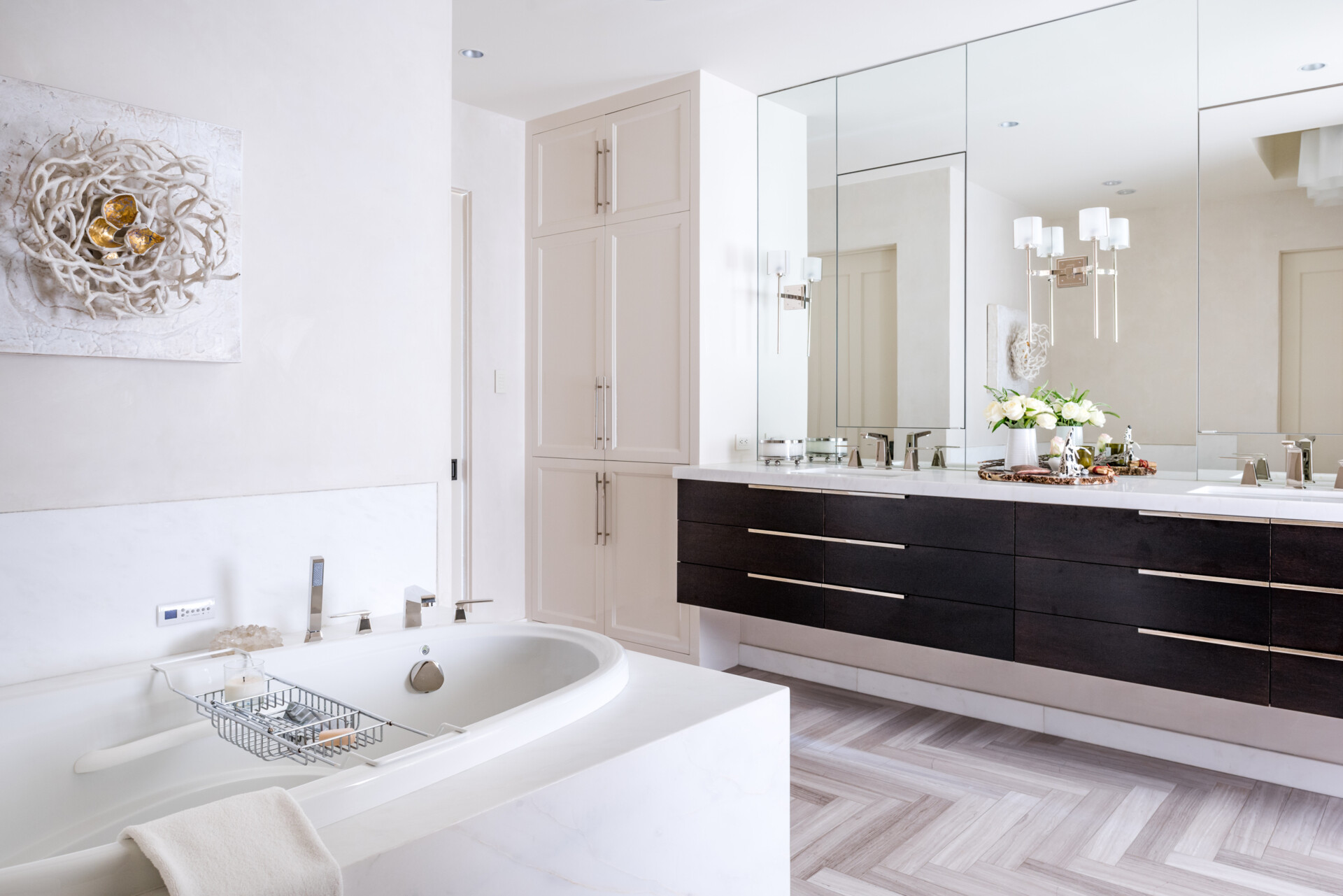 A contemporary bathroom with a built-in bathtub, dark wood floating double vanity, white countertops, large mirrors, herringbone floor tiles, and tall storage cabinets.