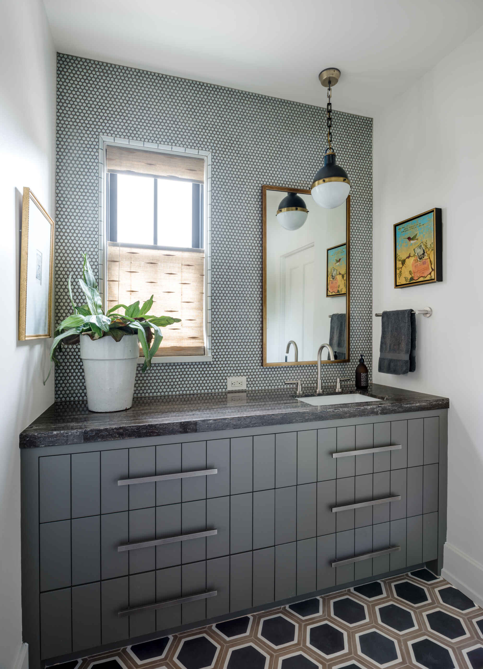 A contemporary bathroom with a gray vanity, dark stone countertop, small circular wall tiles, bold hexagonal floor tiles, and a black-and-white globe pendant light.