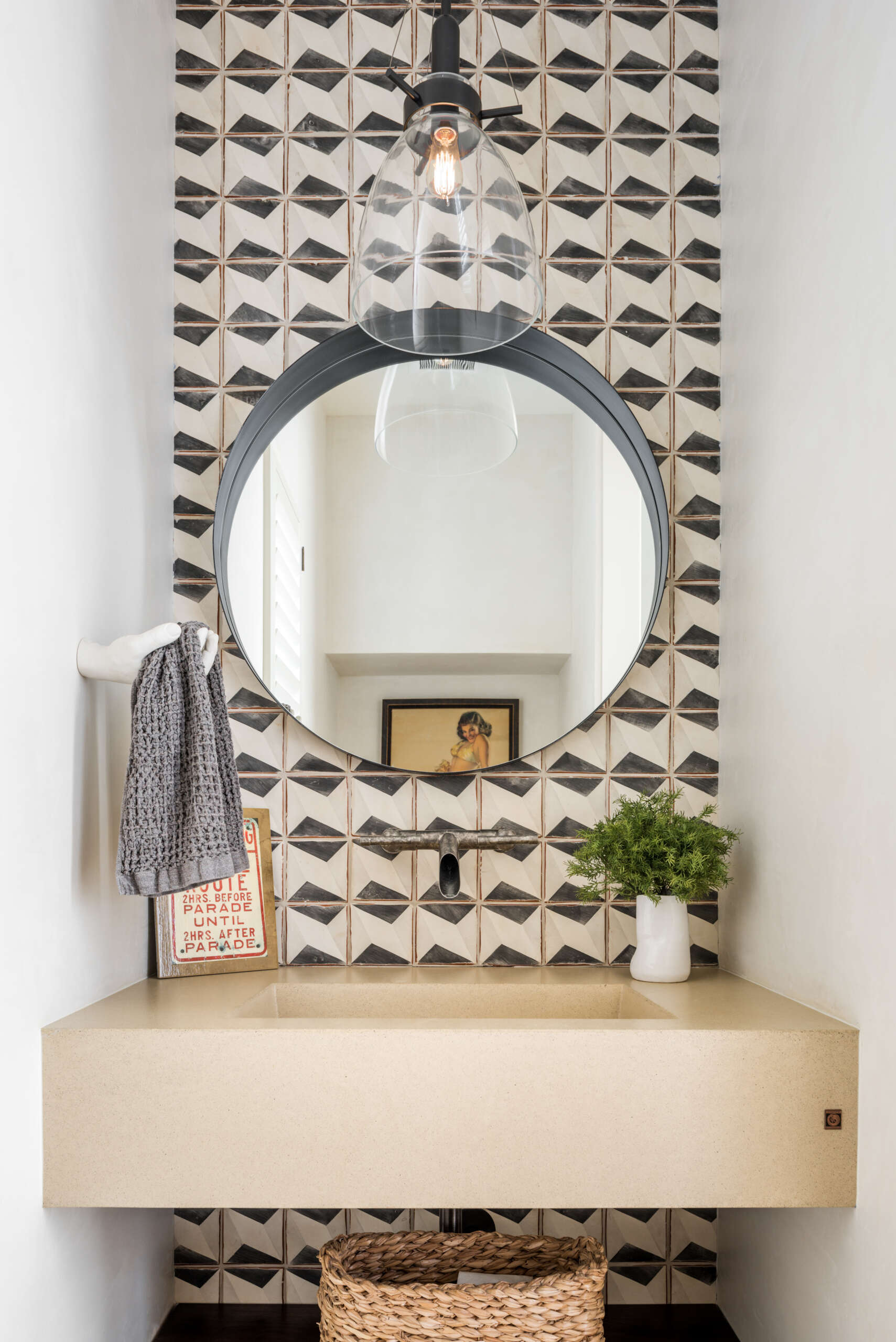 A small contemporary bathroom with black-and-white geometric wall tiles, a floating stone sink, oversized round mirror, wall-mounted faucet, and clear glass pendant lighting.