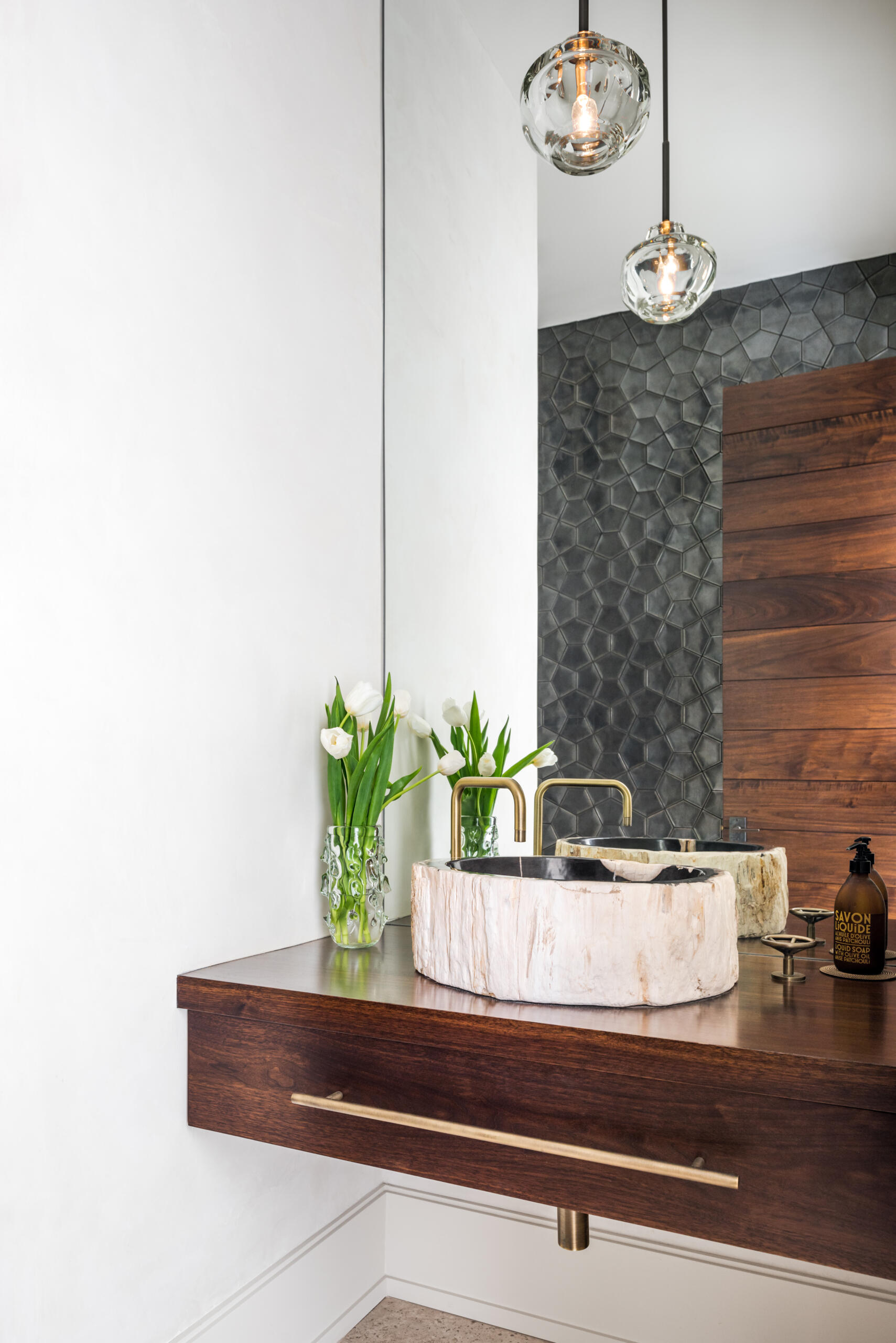 A contemporary bathroom with a dark wood floating vanity, sculptural stone vessel sink, brass faucet, geometric charcoal wall tiles, and clear glass pendant lights.