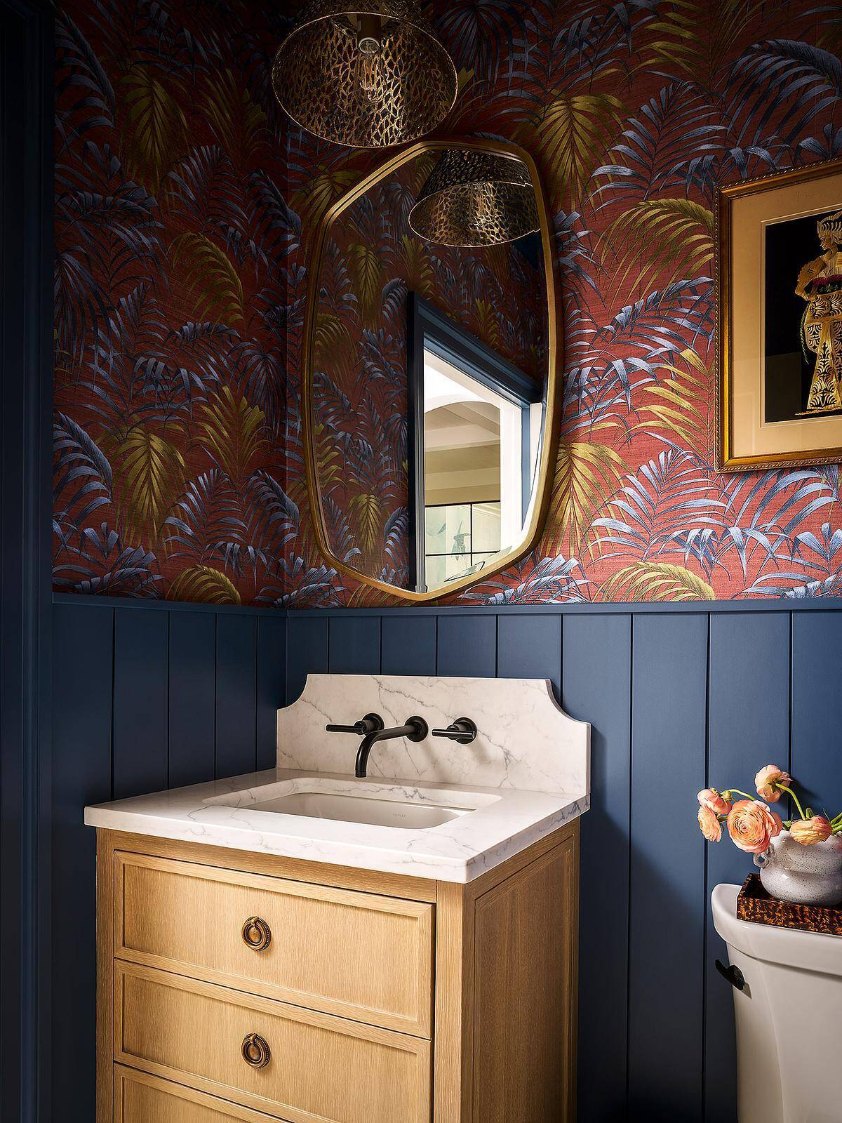 A small bathroom with dramatic red tropical wallpaper, deep navy blue wall paneling, a light wood vanity, marble countertop, black wall-mounted faucet, and an irregularly shaped brass-framed mirror.