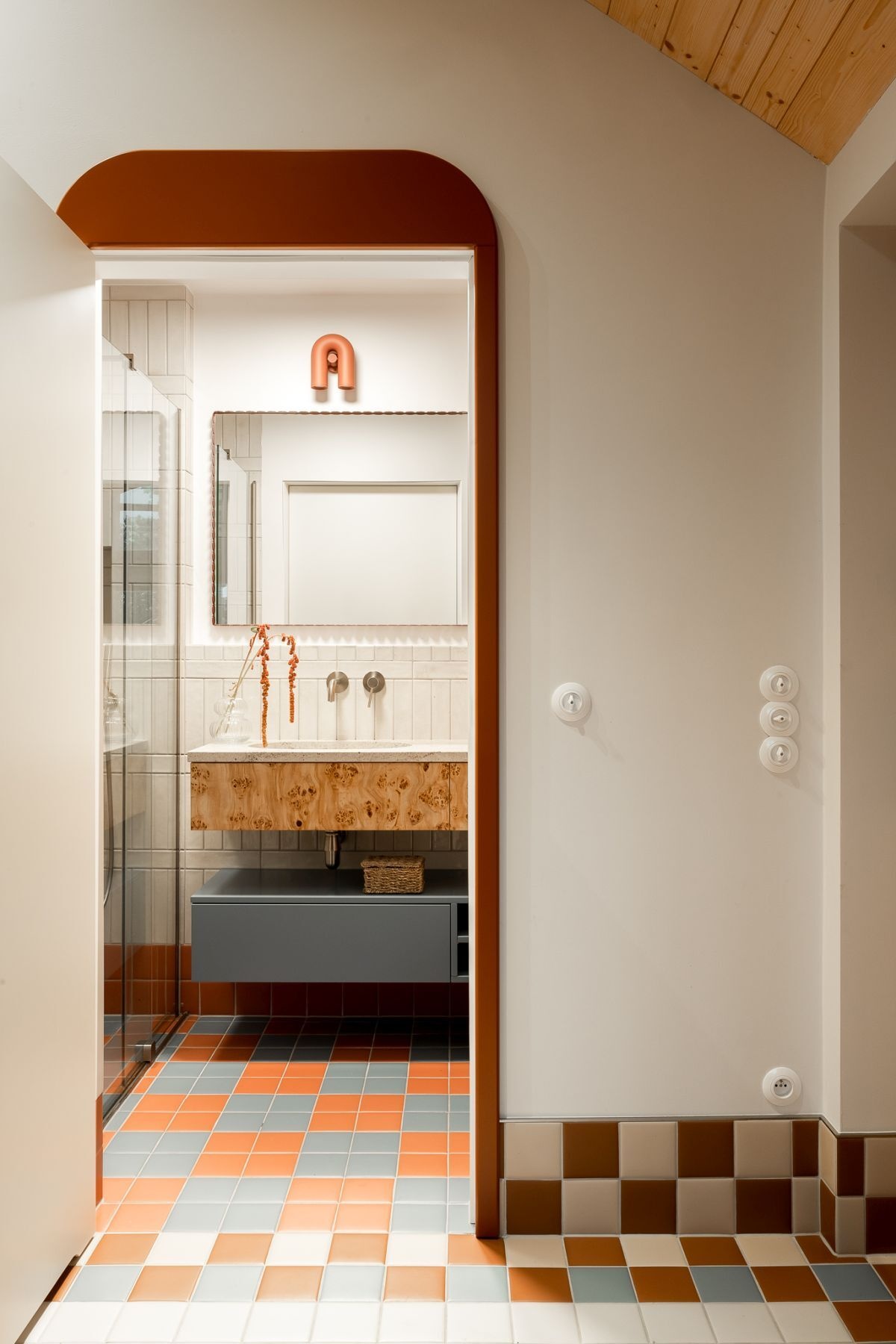 Colorful contemporary bathroom design featuring orange and blue mosaic floor tiles, cream vertical wall tiles, a custom poplar burl wood vanity, scalloped mirror, and terracotta wall lighting.