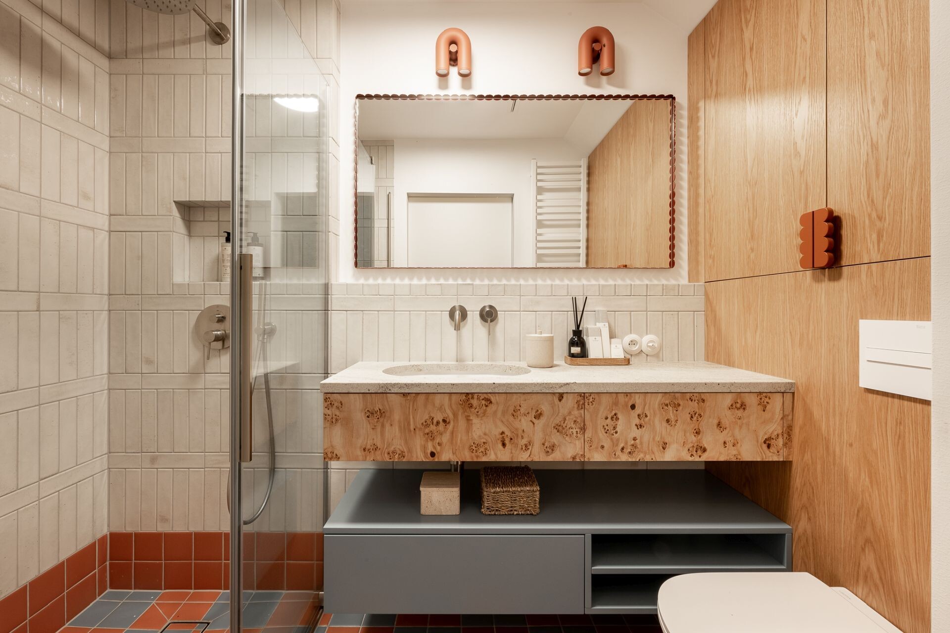Colorful contemporary bathroom design featuring orange and blue mosaic floor tiles, cream vertical wall tiles, a custom poplar burl wood vanity, scalloped mirror, and terracotta wall lighting.