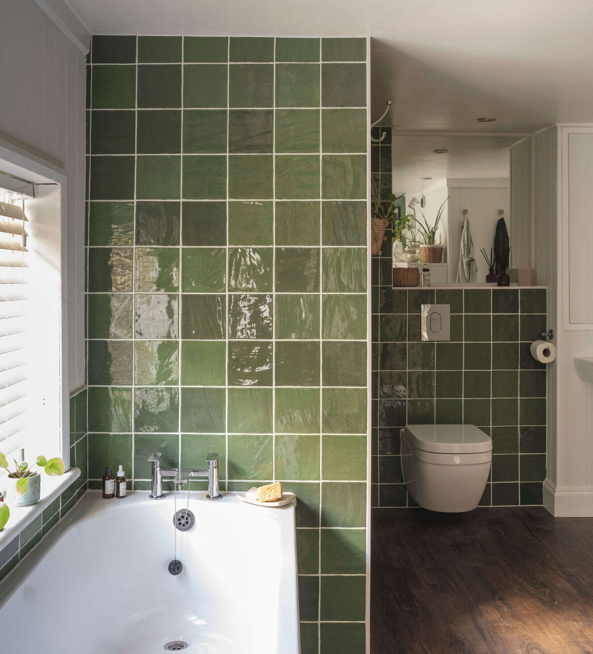 A cottage bathroom with glossy green square tiles, white grout, a bathtub beneath a window, dark flooring, and vertical wall paneling offers colorful renovation inspiration.