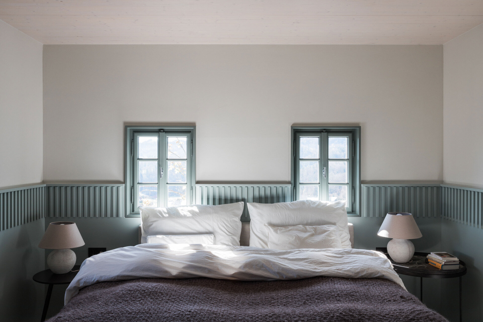 Soft Blue-Grey Panelling and Natural Textures
The ground-floor bedroom introduces a gentle blue-grey colour scheme that gives the room its own identity. A band of vertically grooved panelling wraps around the walls at bed height, continuing between the two small windows positioned above the headboard. The panelling forms a subtle frame for the bed and gives the otherwise plain walls additional texture. Matching window frames extend the colour scheme, while simple bedside tables and rounded ceramic lamps provide a pleasing sense of symmetry.
The bedding introduces a contrasting mix of textures, with loosely arranged white linen pillows and a thick, knitted blanket in a muted purple-grey shade. The room is furnished simply, allowing the colour and architectural detailing to take precedence. A timber ceiling beam adds a reminder of the house's age, while the doorway to the adjoining bathroom offers a glimpse of a completely different material palette.