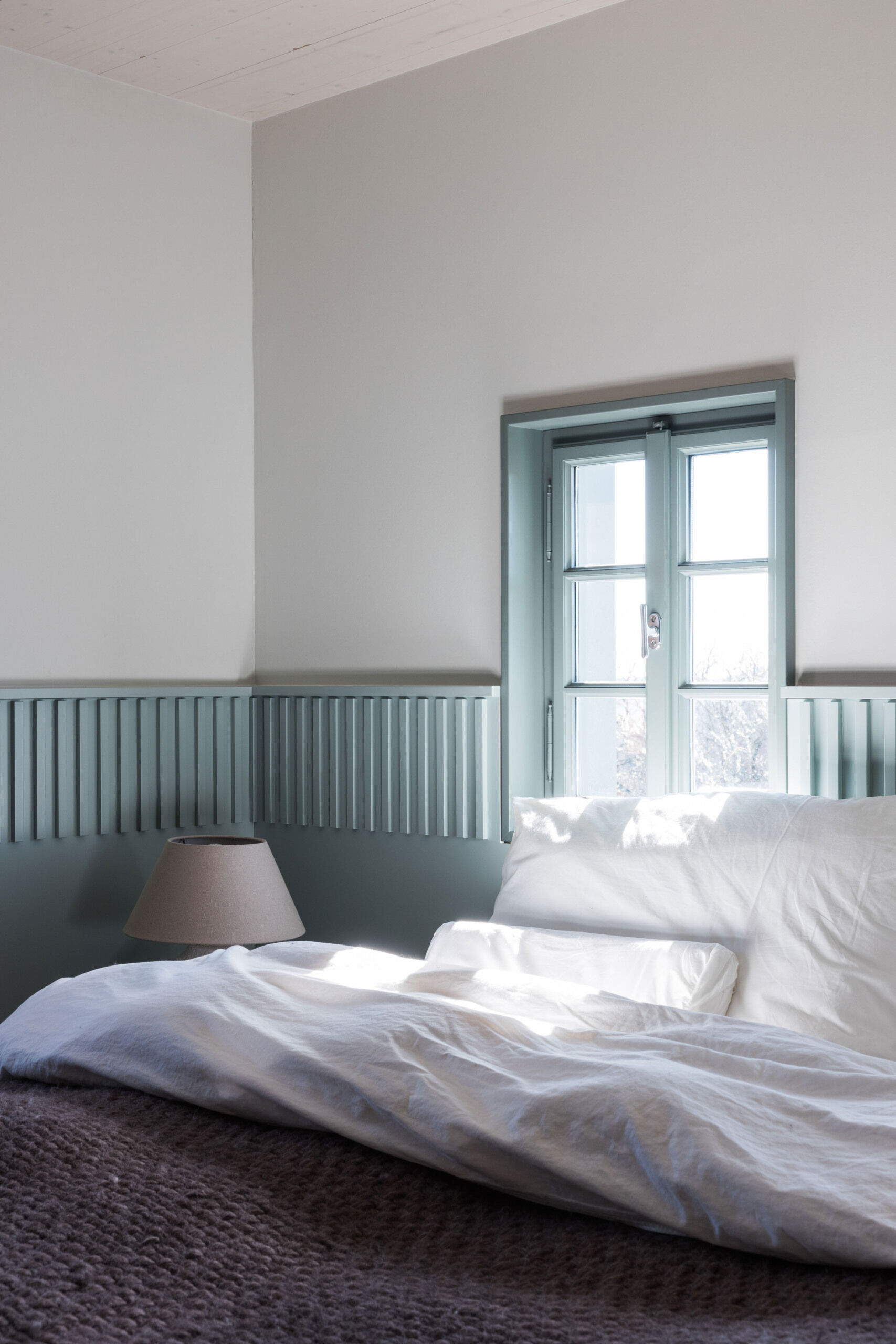 Soft Blue-Grey Panelling and Natural Textures
The ground-floor bedroom introduces a gentle blue-grey colour scheme that gives the room its own identity. A band of vertically grooved panelling wraps around the walls at bed height, continuing between the two small windows positioned above the headboard. The panelling forms a subtle frame for the bed and gives the otherwise plain walls additional texture. Matching window frames extend the colour scheme, while simple bedside tables and rounded ceramic lamps provide a pleasing sense of symmetry.
The bedding introduces a contrasting mix of textures, with loosely arranged white linen pillows and a thick, knitted blanket in a muted purple-grey shade. The room is furnished simply, allowing the colour and architectural detailing to take precedence. A timber ceiling beam adds a reminder of the house's age, while the doorway to the adjoining bathroom offers a glimpse of a completely different material palette.