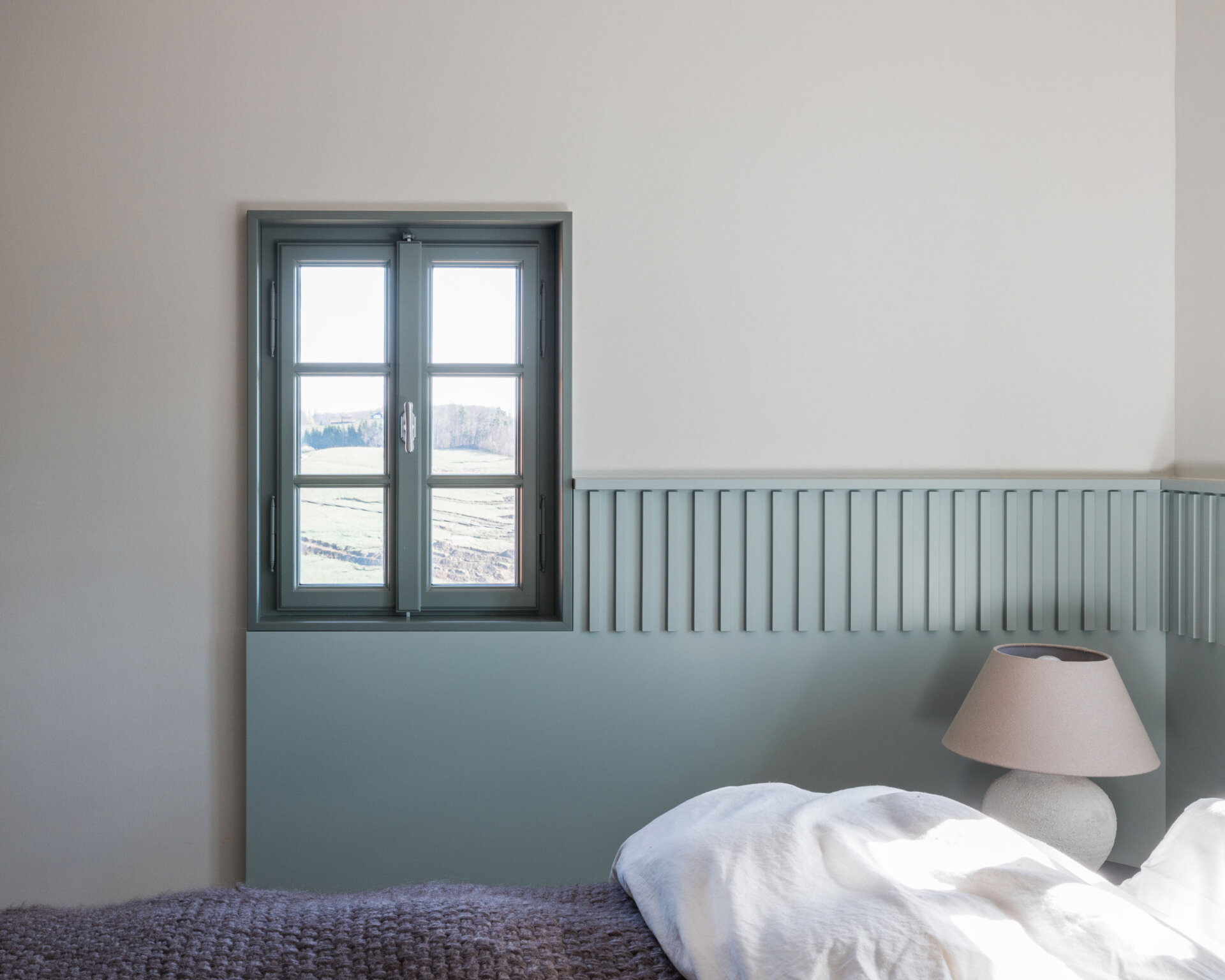Soft Blue-Grey Panelling and Natural Textures
The ground-floor bedroom introduces a gentle blue-grey colour scheme that gives the room its own identity. A band of vertically grooved panelling wraps around the walls at bed height, continuing between the two small windows positioned above the headboard. The panelling forms a subtle frame for the bed and gives the otherwise plain walls additional texture. Matching window frames extend the colour scheme, while simple bedside tables and rounded ceramic lamps provide a pleasing sense of symmetry.
The bedding introduces a contrasting mix of textures, with loosely arranged white linen pillows and a thick, knitted blanket in a muted purple-grey shade. The room is furnished simply, allowing the colour and architectural detailing to take precedence. A timber ceiling beam adds a reminder of the house's age, while the doorway to the adjoining bathroom offers a glimpse of a completely different material palette.