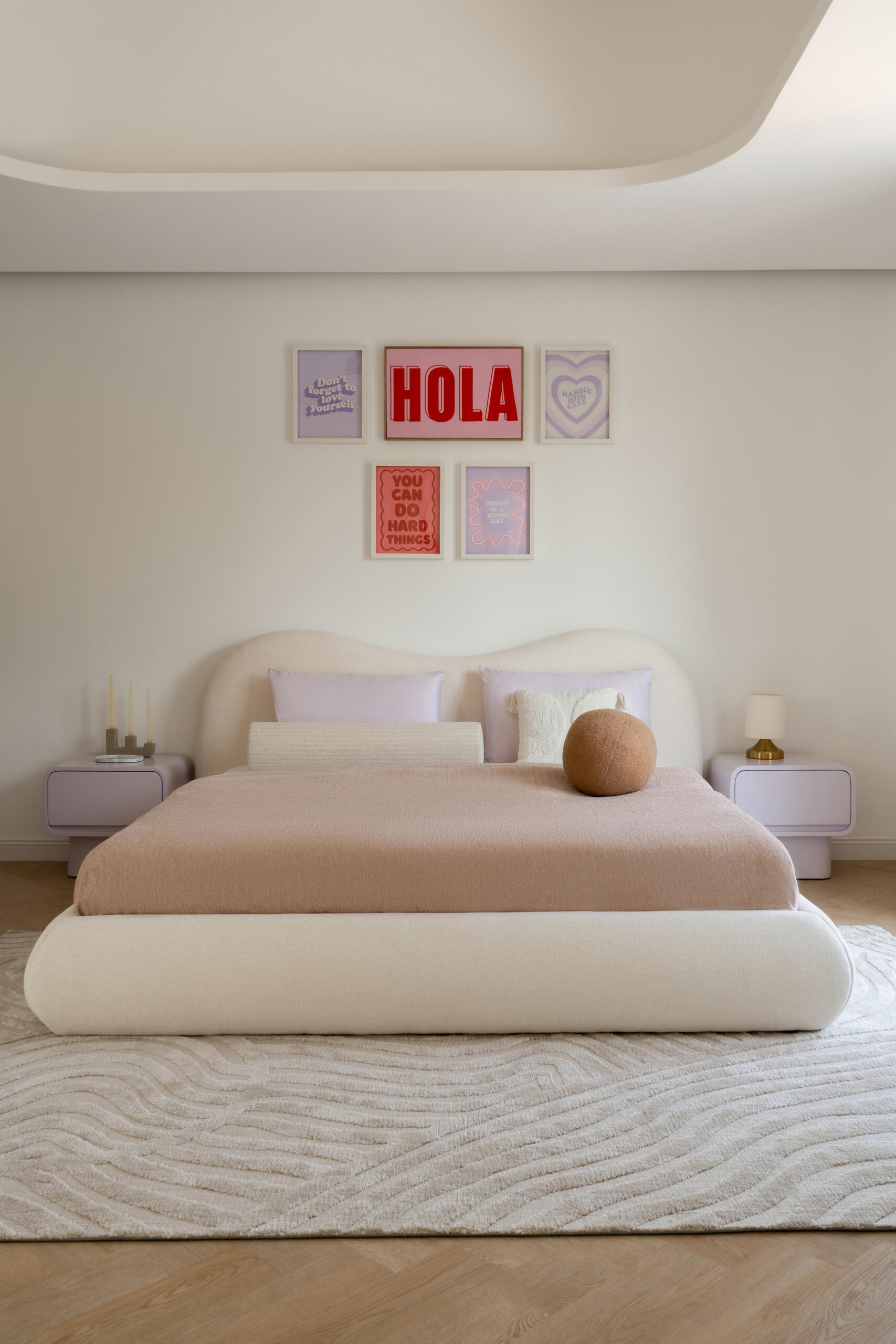 A contemporary bedroom inside The White Arcade in Dubai. Designed by D’Ora Tokai Designs, this youthful bedroom features a custom curved upholstered bed, pastel lavender bedside tables, colourful wall art, a textured white rug, and an adjoining dressing area.