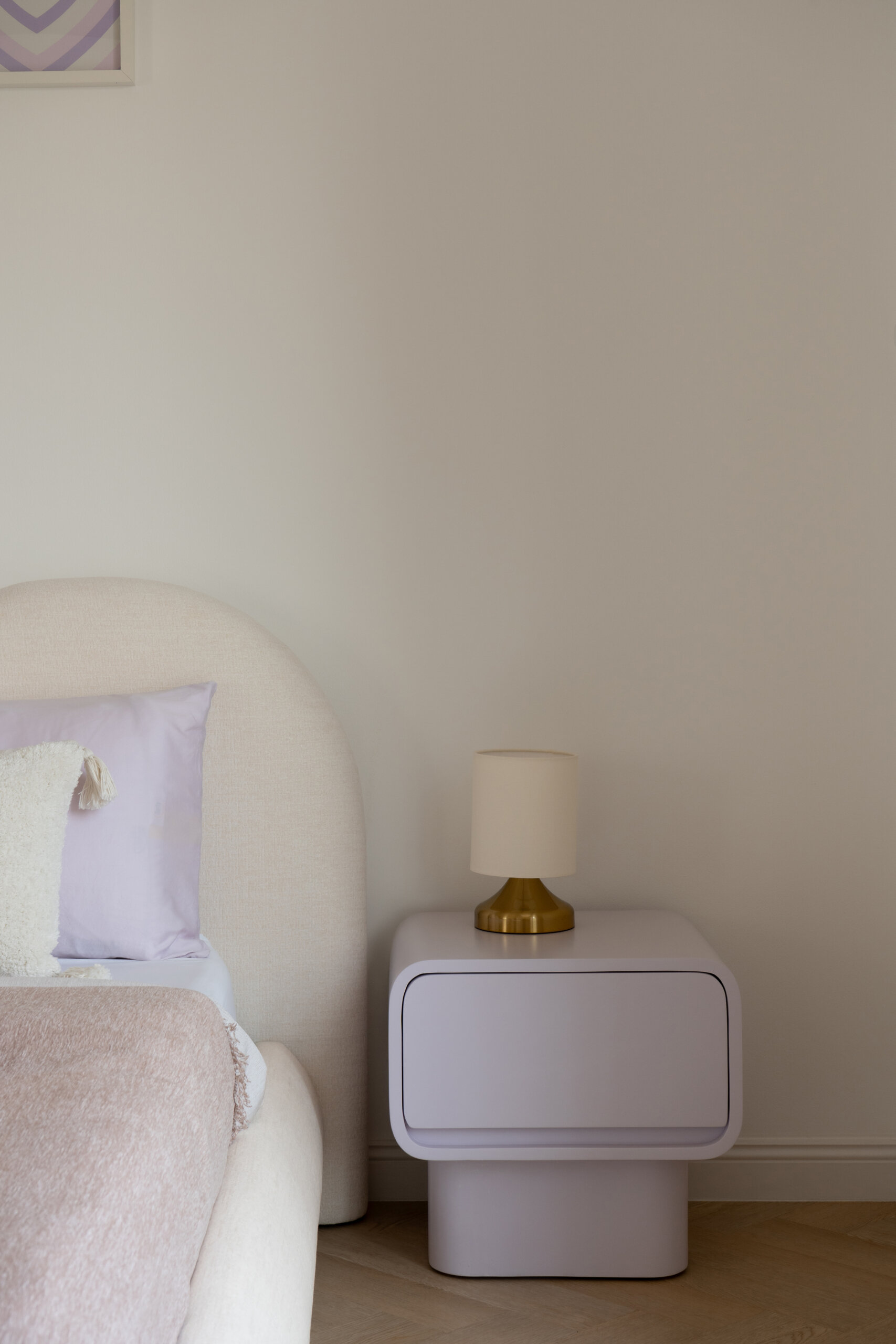 A contemporary bedroom inside The White Arcade in Dubai. Designed by D’Ora Tokai Designs, this youthful bedroom features a custom curved upholstered bed, pastel lavender bedside tables, colourful wall art, a textured white rug, and an adjoining dressing area.