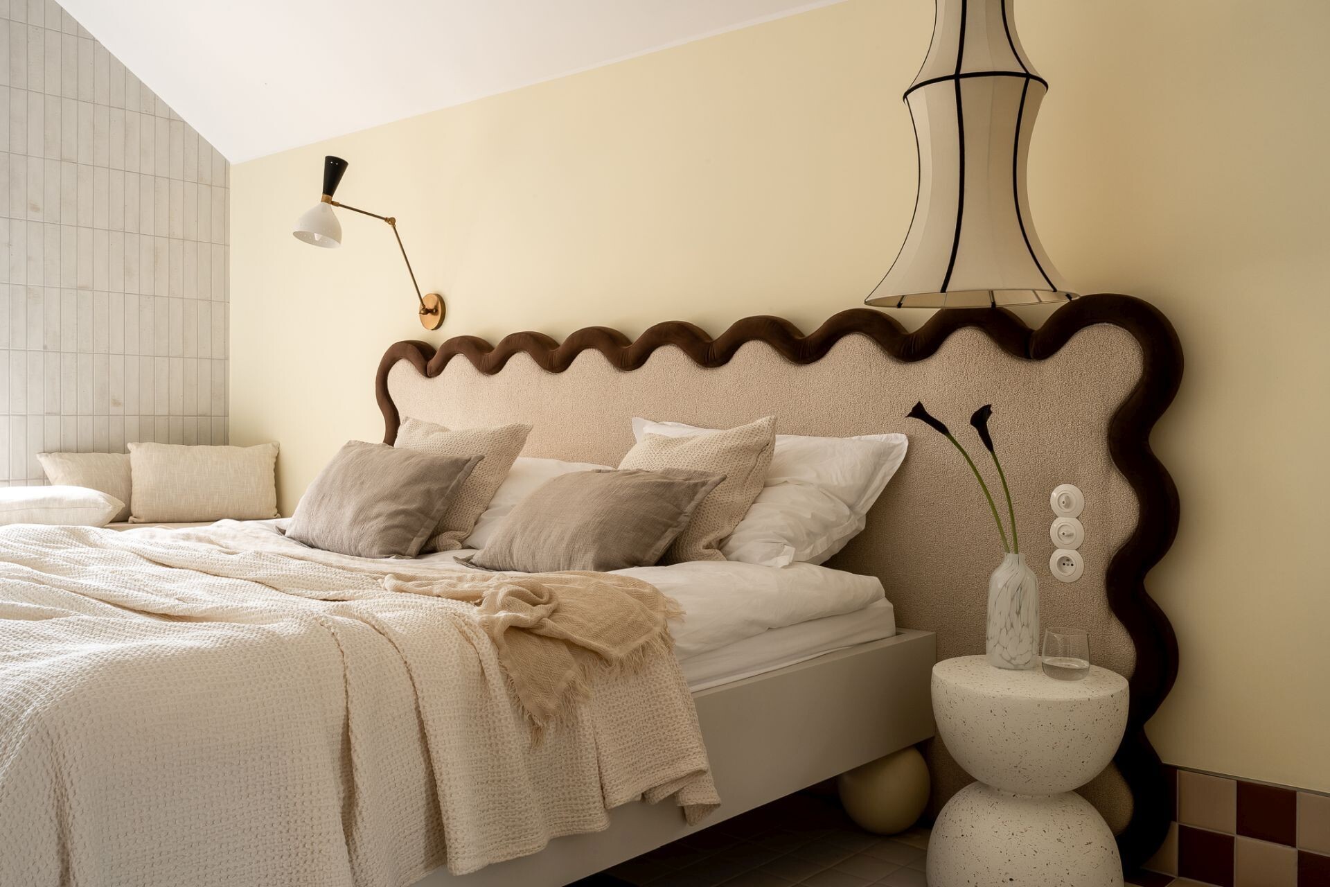 Bedroom design inspiration featuring a sculptural wave-shaped headboard, neutral bedding, unusual pendant lighting, custom fitted wardrobes, and bold black-and-white wallpaper.