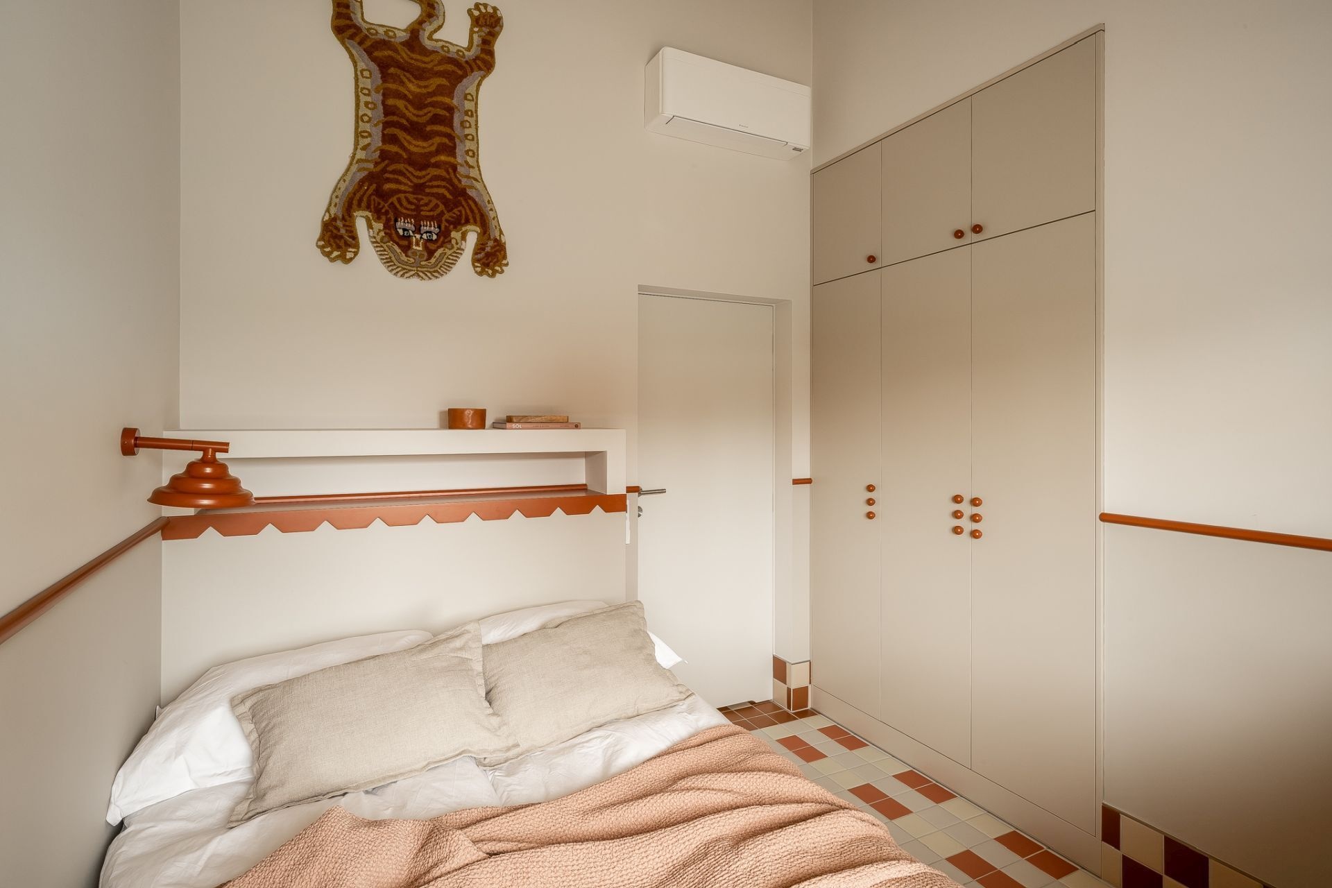 A small contemporary bedroom featuring cream built-in wardrobes, terracotta accents, a custom headboard with zigzag trim, soft pink bedding, and a playful tiger wall hanging.