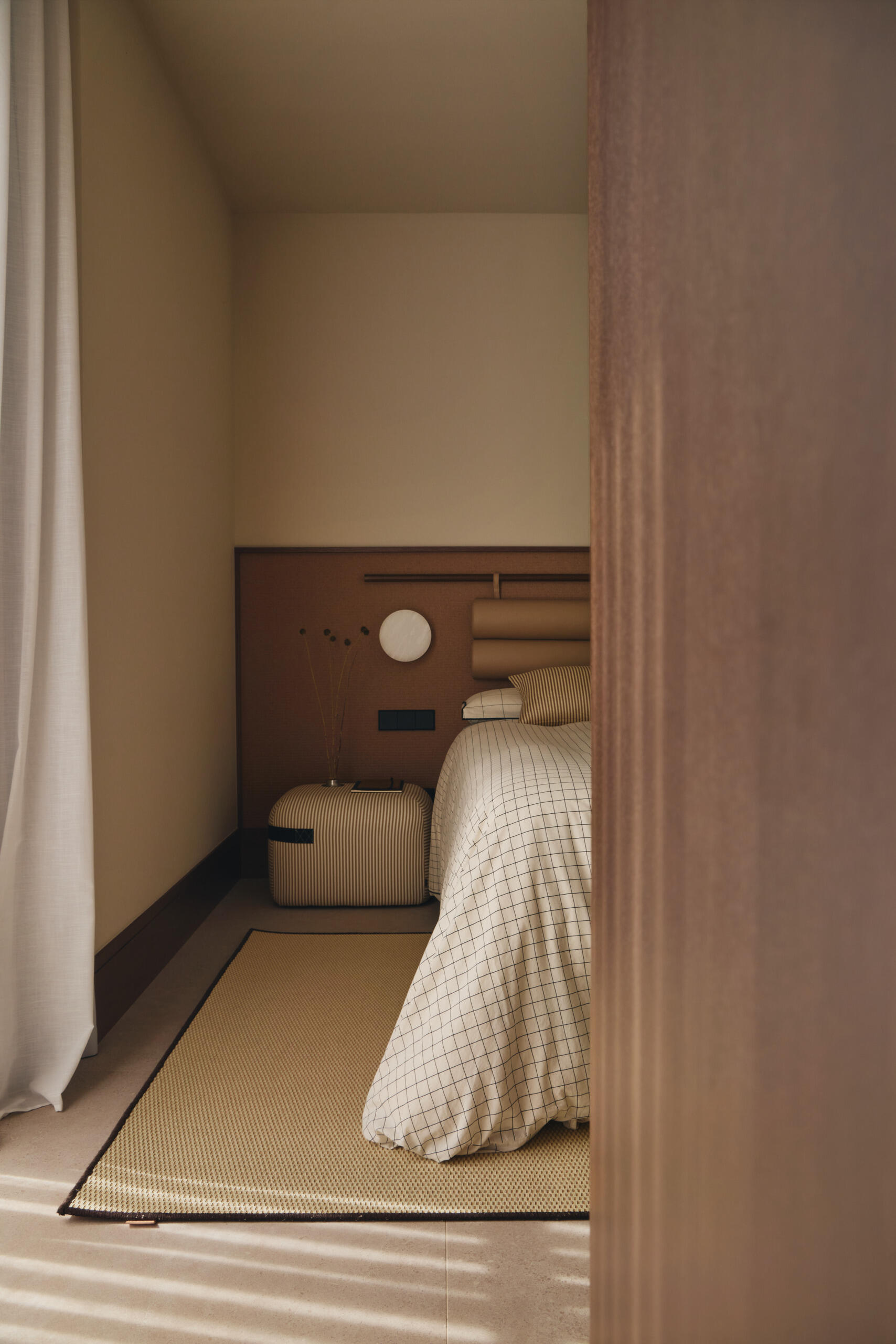 Discover a contemporary bedroom with a woven wall headboard, neutral checked bedding, striped bedside pouf, and custom timber desk integrated into the room's cabinetry.