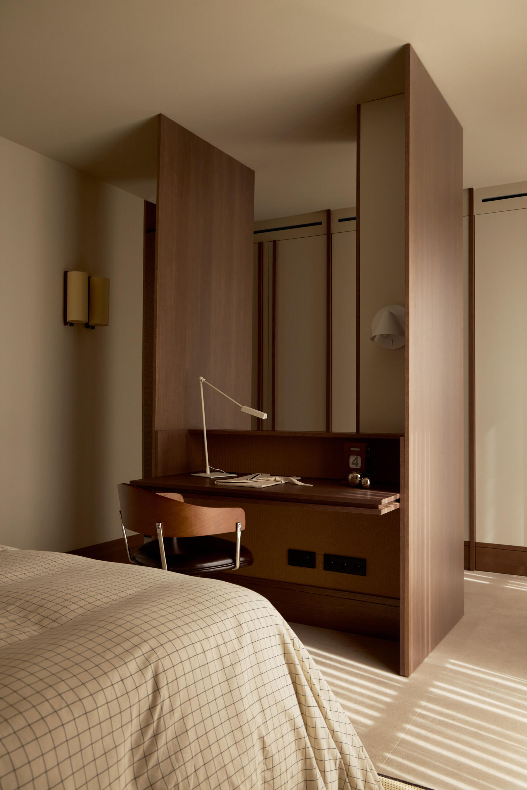 Discover a contemporary bedroom with a woven wall headboard, neutral checked bedding, striped bedside pouf, and custom timber desk integrated into the room's cabinetry.