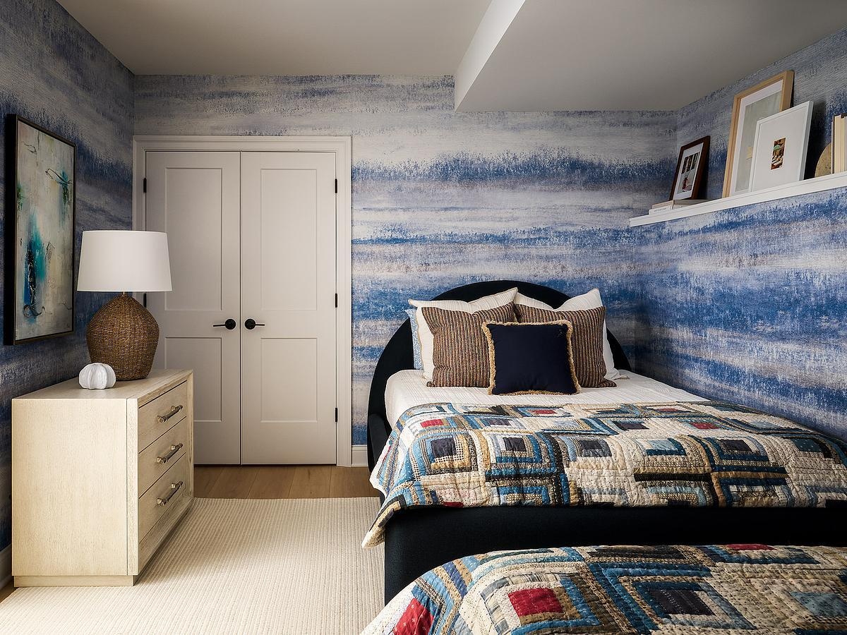 A blue bedroom featuring textured striped wallpaper, geometric patchwork quilts, dark curved headboards, a light-colored dresser, and a woven table lamp.