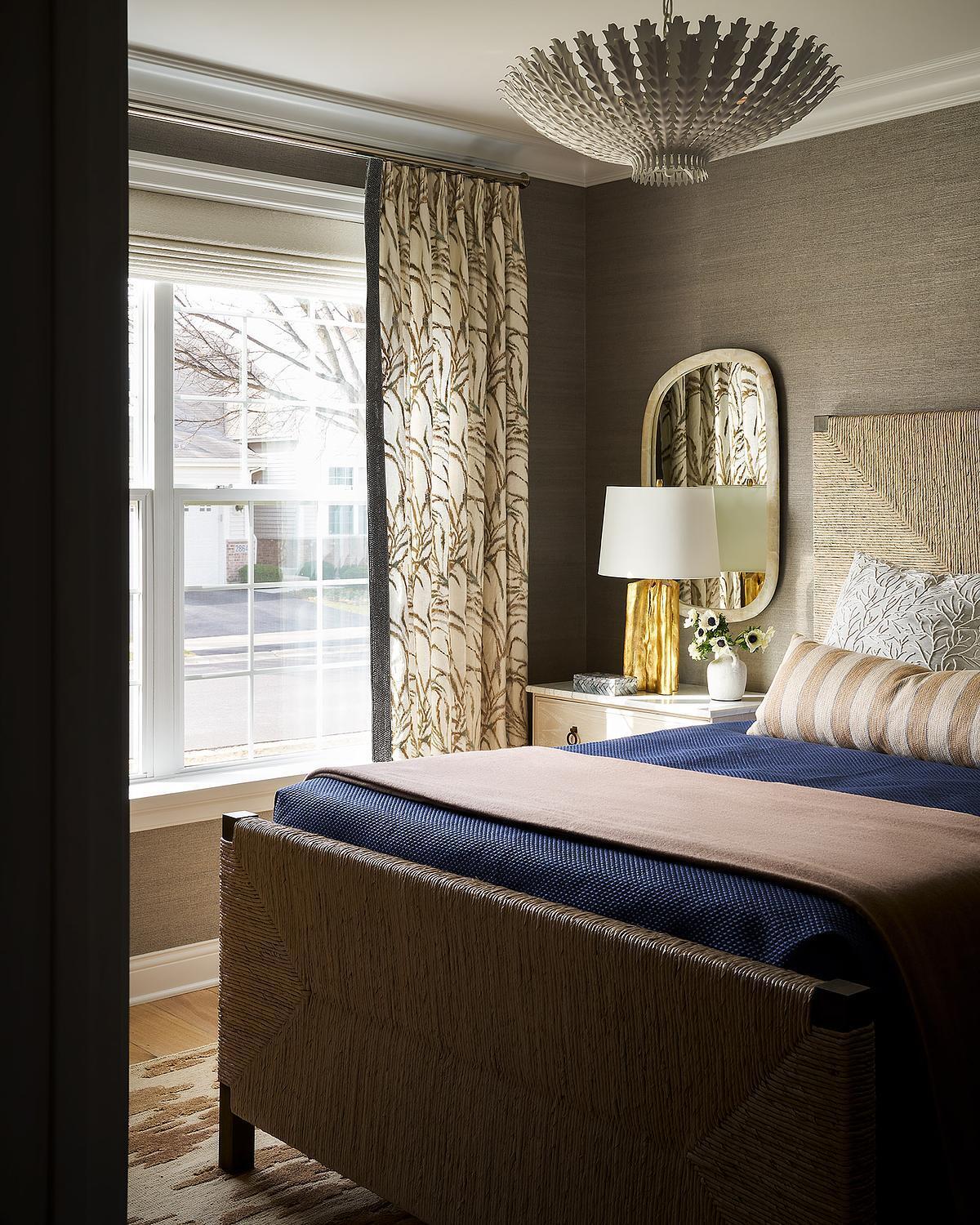 A contemporary bedroom with deep blue bedding, a textured upholstered bed frame, botanical patterned curtains, a gold table lamp, and a neutral woven wall covering.