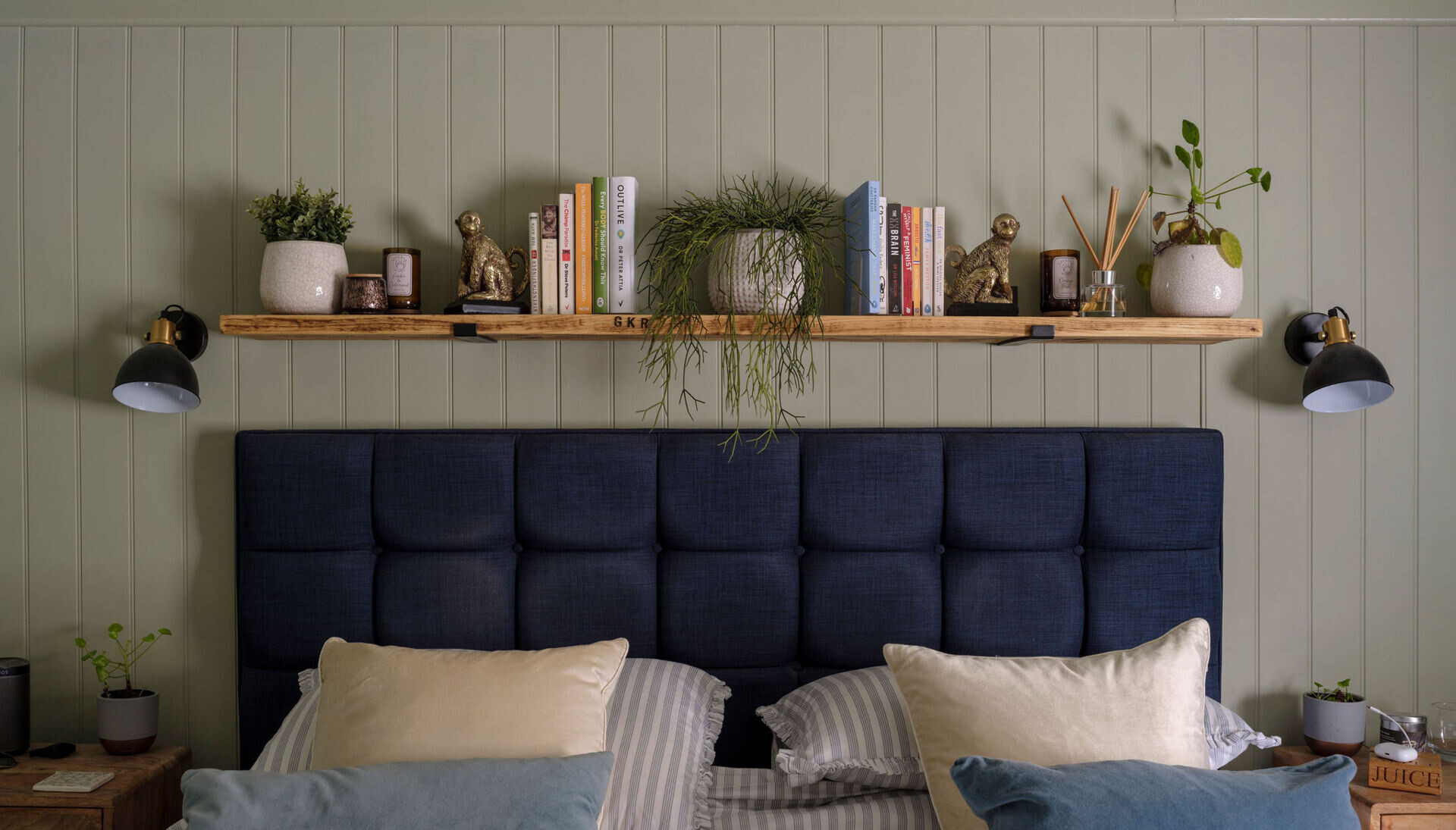 A cozy cottage bedroom features sage-green wall paneling, a navy upholstered headboard, floating wood shelving, and matching black-and-brass bedside lights.