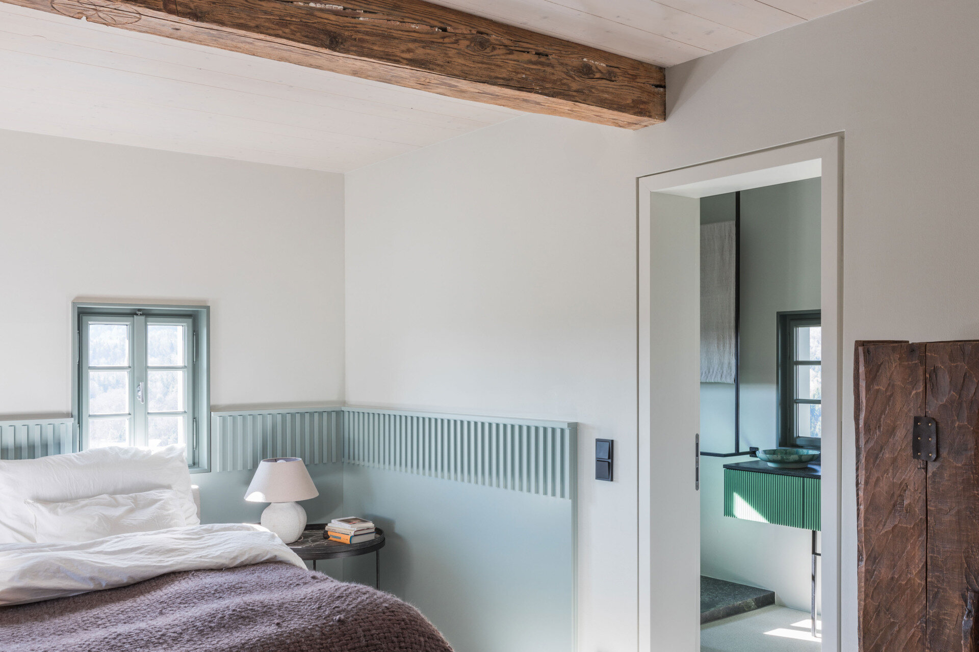 A contemporary ensuite bathroom with dark stone walls, a glass shower enclosure, green fluted vanity, ceramic vessel basin, and unusual architectural detailing.
The ground-floor bedroom introduces a gentle blue-grey colour scheme that gives the room its own identity. A band of vertically grooved panelling wraps around the walls at bed height, continuing between the two small windows positioned above the headboard. The panelling forms a subtle frame for the bed and gives the otherwise plain walls additional texture. Matching window frames extend the colour scheme, while simple bedside tables and rounded ceramic lamps provide a pleasing sense of symmetry.
The bedding introduces a contrasting mix of textures, with loosely arranged white linen pillows and a thick, knitted blanket in a muted purple-grey shade. The room is furnished simply, allowing the colour and architectural detailing to take precedence. A timber ceiling beam adds a reminder of the house's age, while the doorway to the adjoining bathroom offers a glimpse of a completely different material palette.