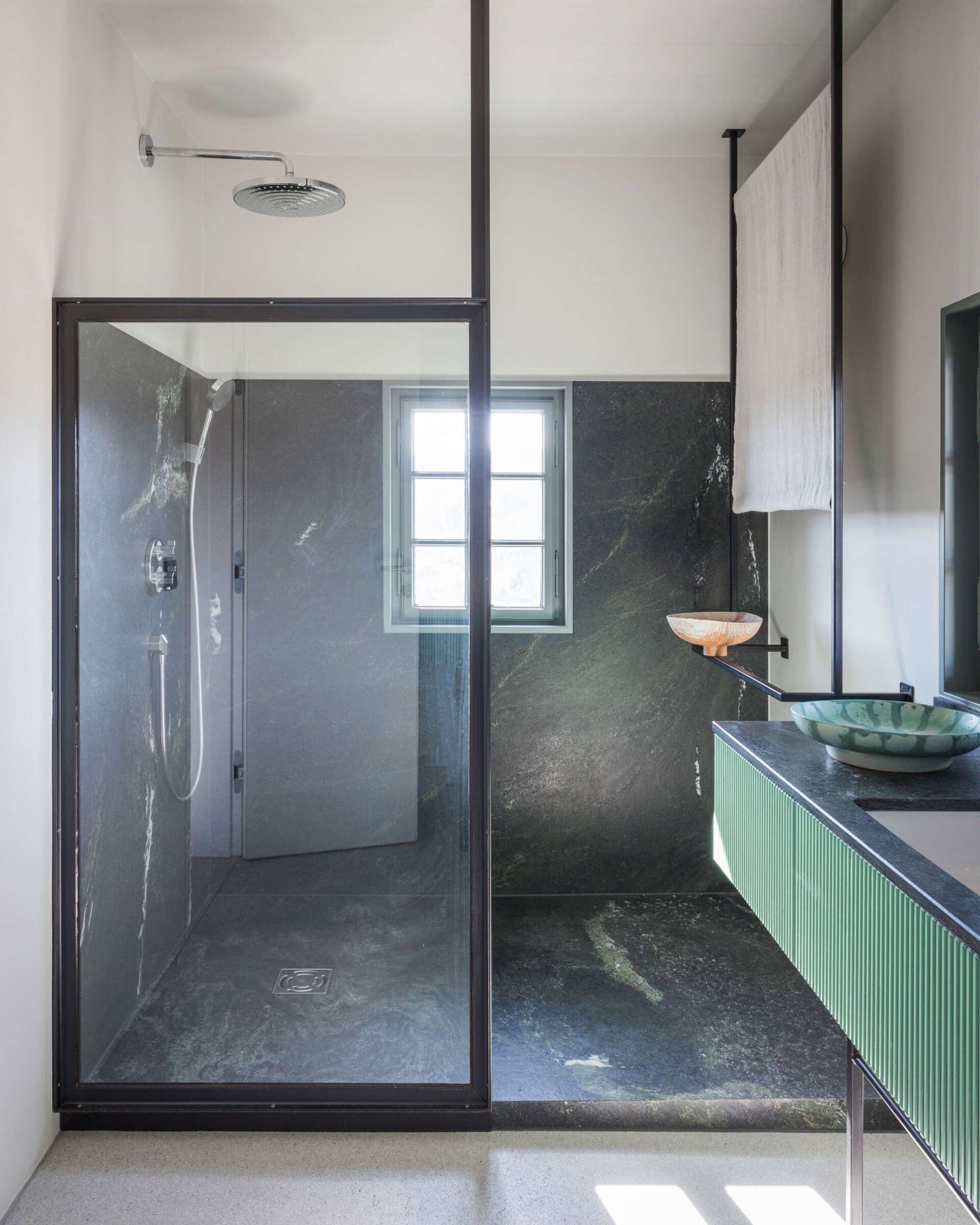 A contemporary ensuite bathroom with dark stone walls, a glass shower enclosure, green fluted vanity, ceramic vessel basin, and unusual architectural detailing.