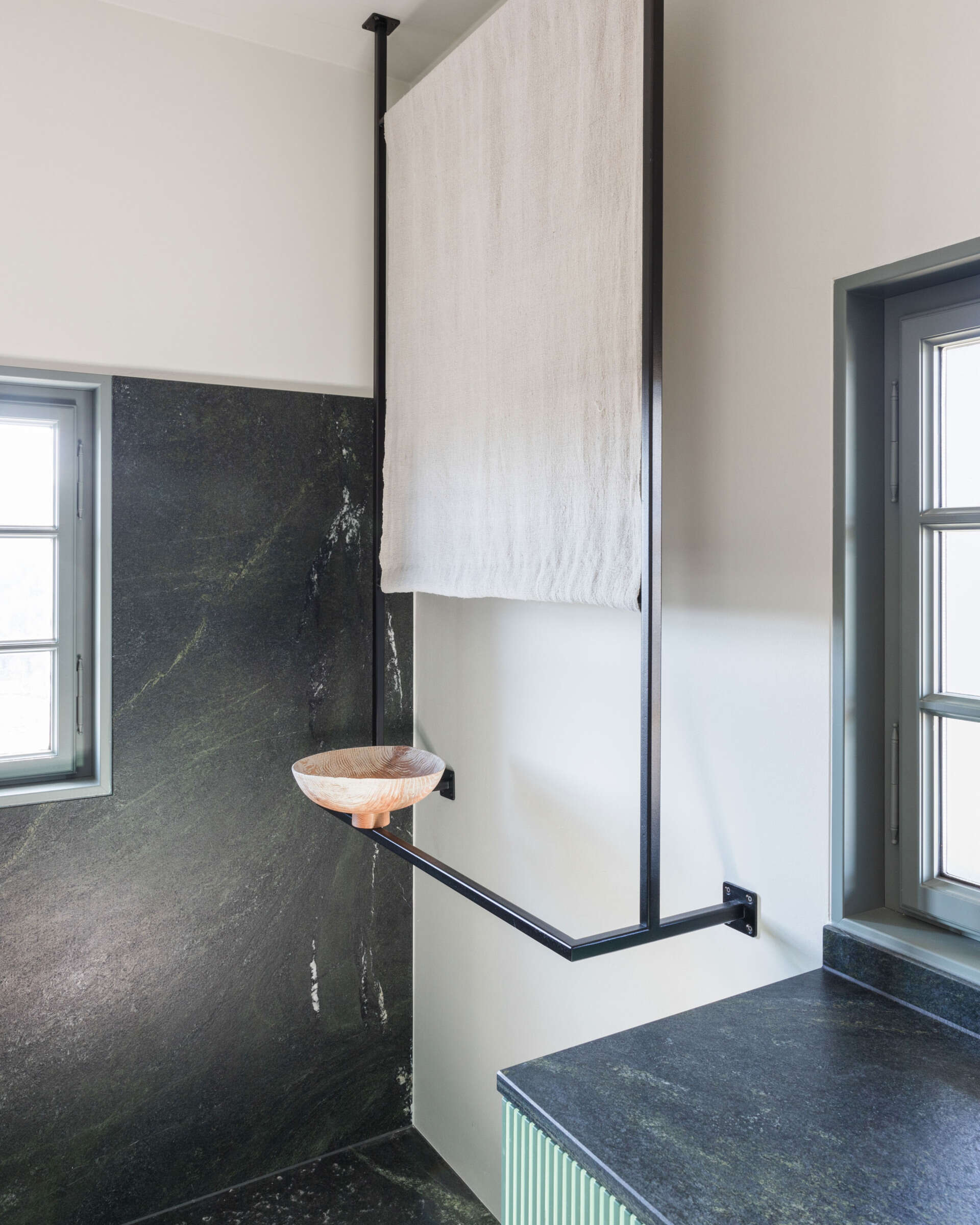 A contemporary ensuite bathroom with dark stone walls, a glass shower enclosure, green fluted vanity, ceramic vessel basin, and unusual architectural detailing.