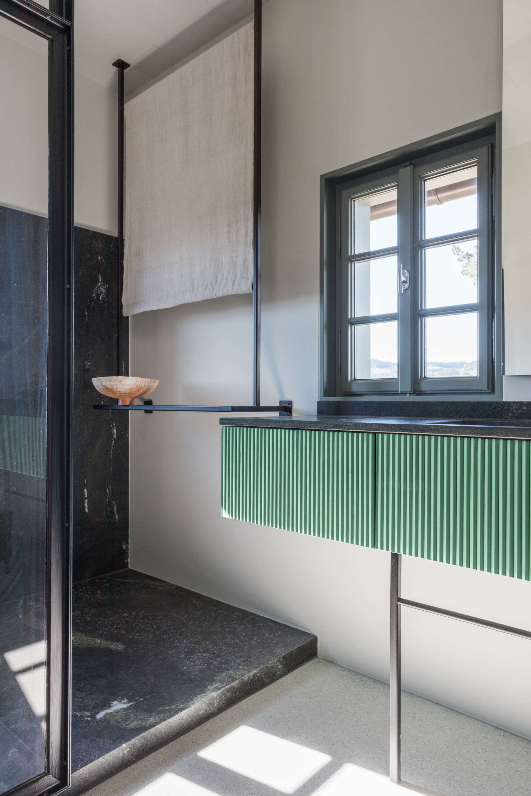 A contemporary ensuite bathroom with dark stone walls, a glass shower enclosure, green fluted vanity, ceramic vessel basin, and unusual architectural detailing.