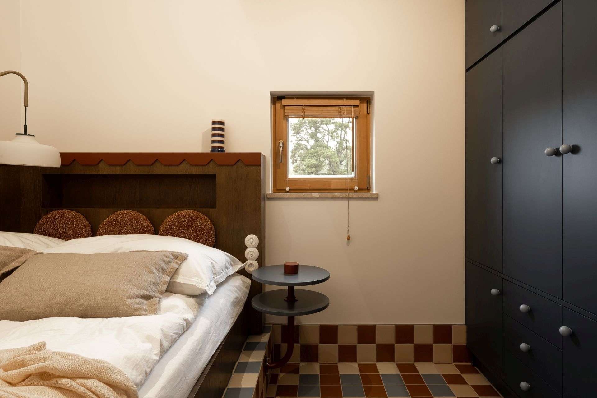 Contemporary bedroom featuring a custom wooden bed, dark blue-gray built-in wardrobes, an oval mirror, and a large upholstered window seat overlooking the outdoor swimming pool.