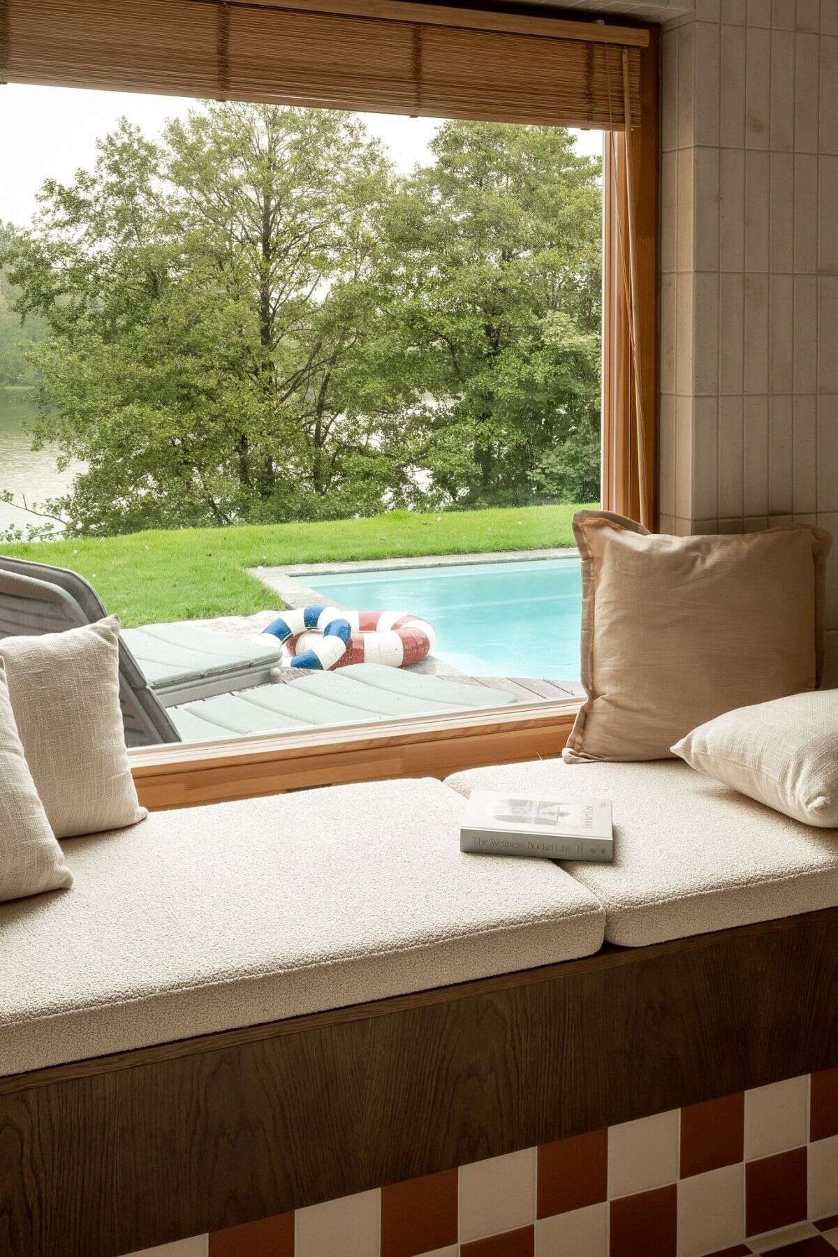 Contemporary bedroom featuring a custom wooden bed, dark blue-gray built-in wardrobes, an oval mirror, and a large upholstered window seat overlooking the outdoor swimming pool.