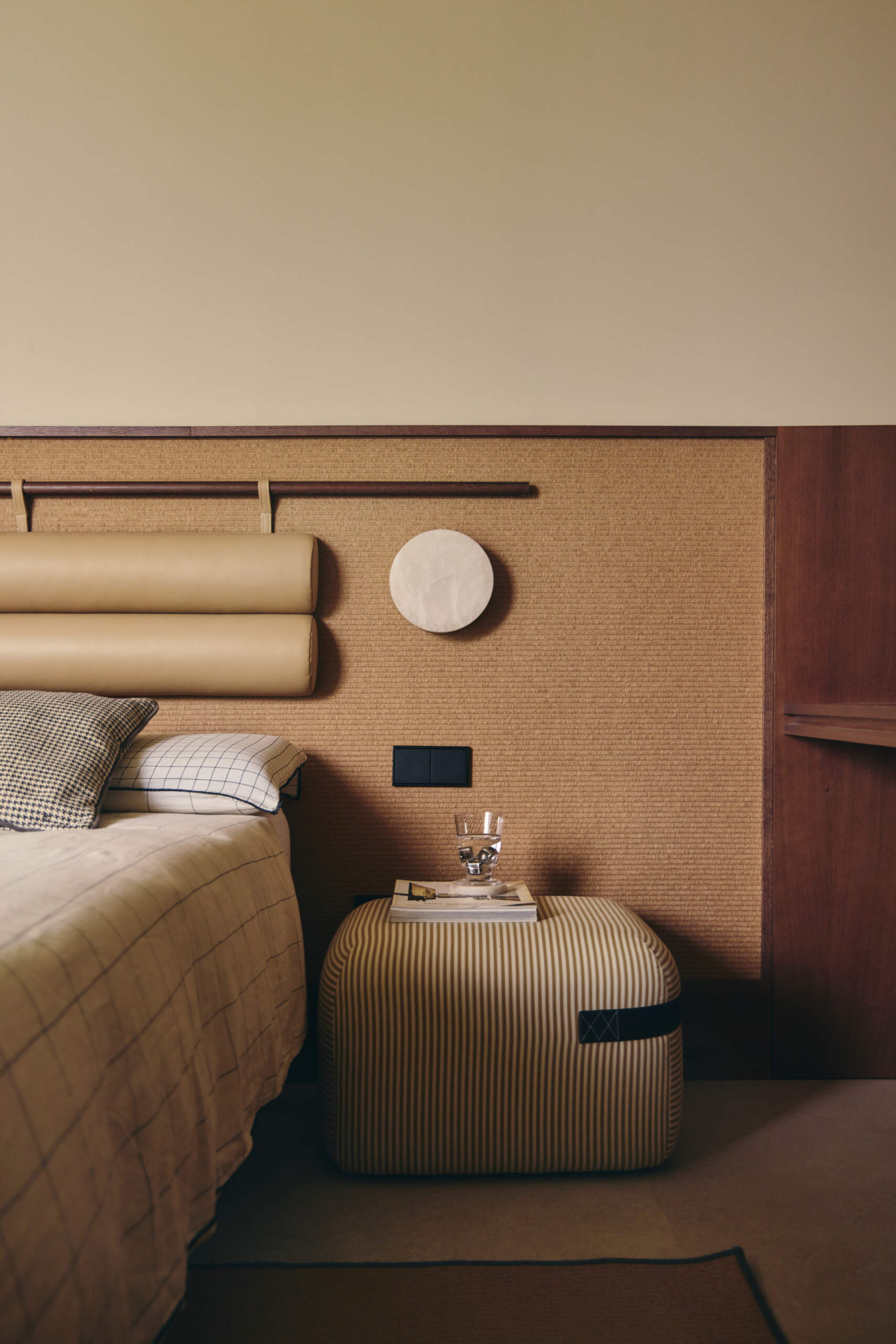 A Second Bedroom with a Built-In Desk and Woven Headboard
The second bedroom follows a similar neutral palette but has a different furniture arrangement. Here, the bed sits against a long, low wall panel that combines a natural woven surface with a timber frame and padded cylindrical headboard. A striped Season pouf by Viccarbe provides a casual alternative to a traditional bedside table, topped with books and a glass of water. The simple bedding includes fine checked patterns and a contrasting blue-and-white cushion.

Along the adjoining wall, a custom timber desk extends beneath an open shelf, creating a generous workspace with integrated storage. A slim metal chair and adjustable white desk lamp keep the arrangement simple, while small cupboards blend into the surrounding cabinetry. The desk is positioned beside the bed, allowing the room to accommodate both sleeping and studying without introducing too many separate pieces of furniture.