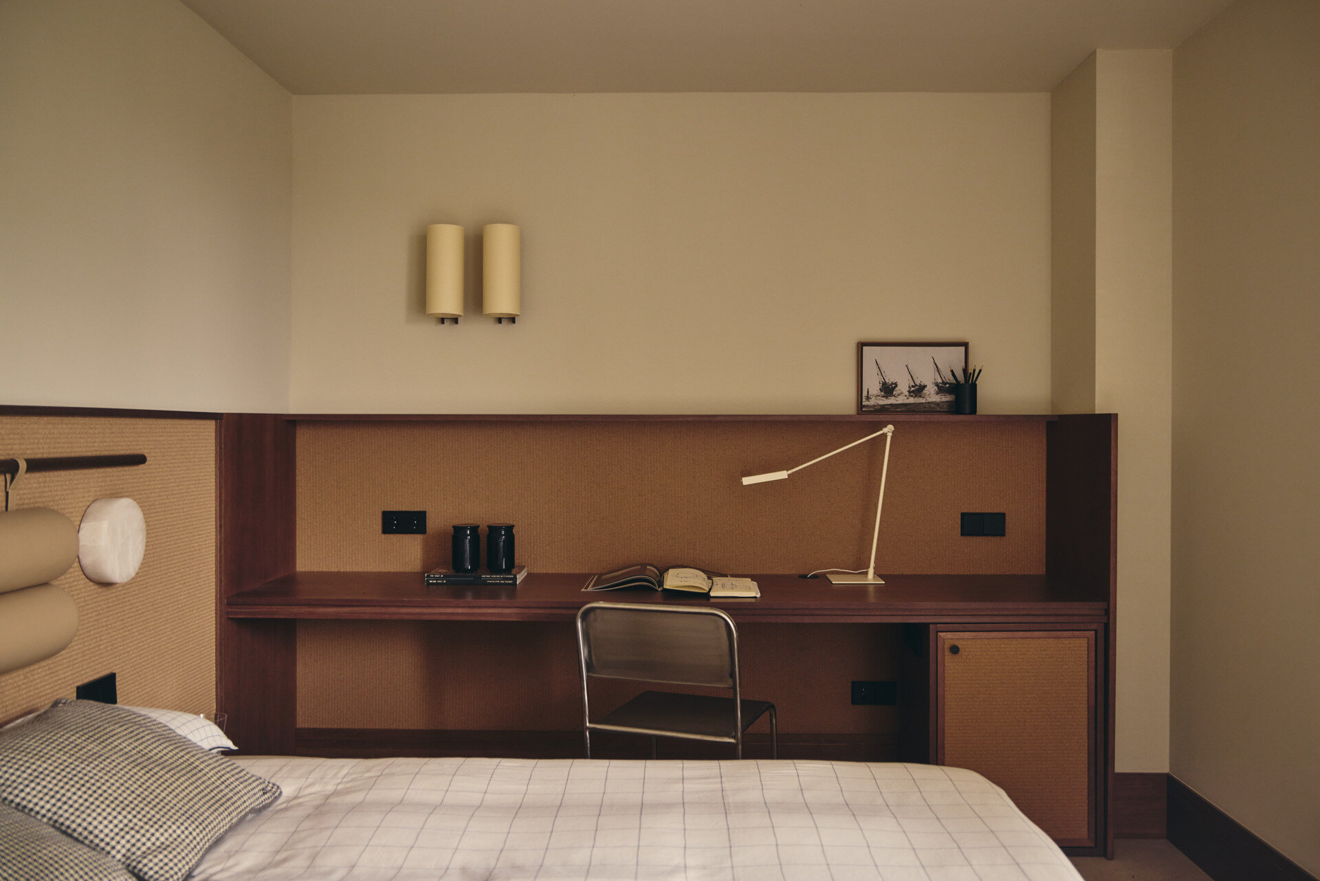 A Second Bedroom with a Built-In Desk and Woven Headboard
The second bedroom follows a similar neutral palette but has a different furniture arrangement. Here, the bed sits against a long, low wall panel that combines a natural woven surface with a timber frame and padded cylindrical headboard. A striped Season pouf by Viccarbe provides a casual alternative to a traditional bedside table, topped with books and a glass of water. The simple bedding includes fine checked patterns and a contrasting blue-and-white cushion.

Along the adjoining wall, a custom timber desk extends beneath an open shelf, creating a generous workspace with integrated storage. A slim metal chair and adjustable white desk lamp keep the arrangement simple, while small cupboards blend into the surrounding cabinetry. The desk is positioned beside the bed, allowing the room to accommodate both sleeping and studying without introducing too many separate pieces of furniture.
