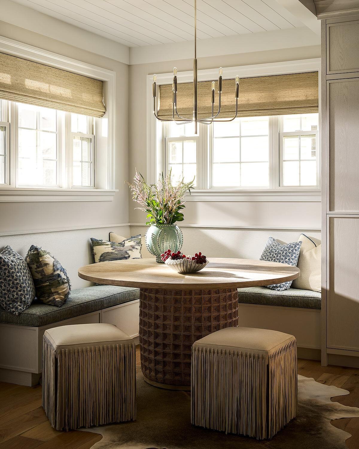 A bright breakfast nook with built-in upholstered banquette seating, a round wood dining table with a textured pedestal base, fringed stools, and windows with woven shades.
