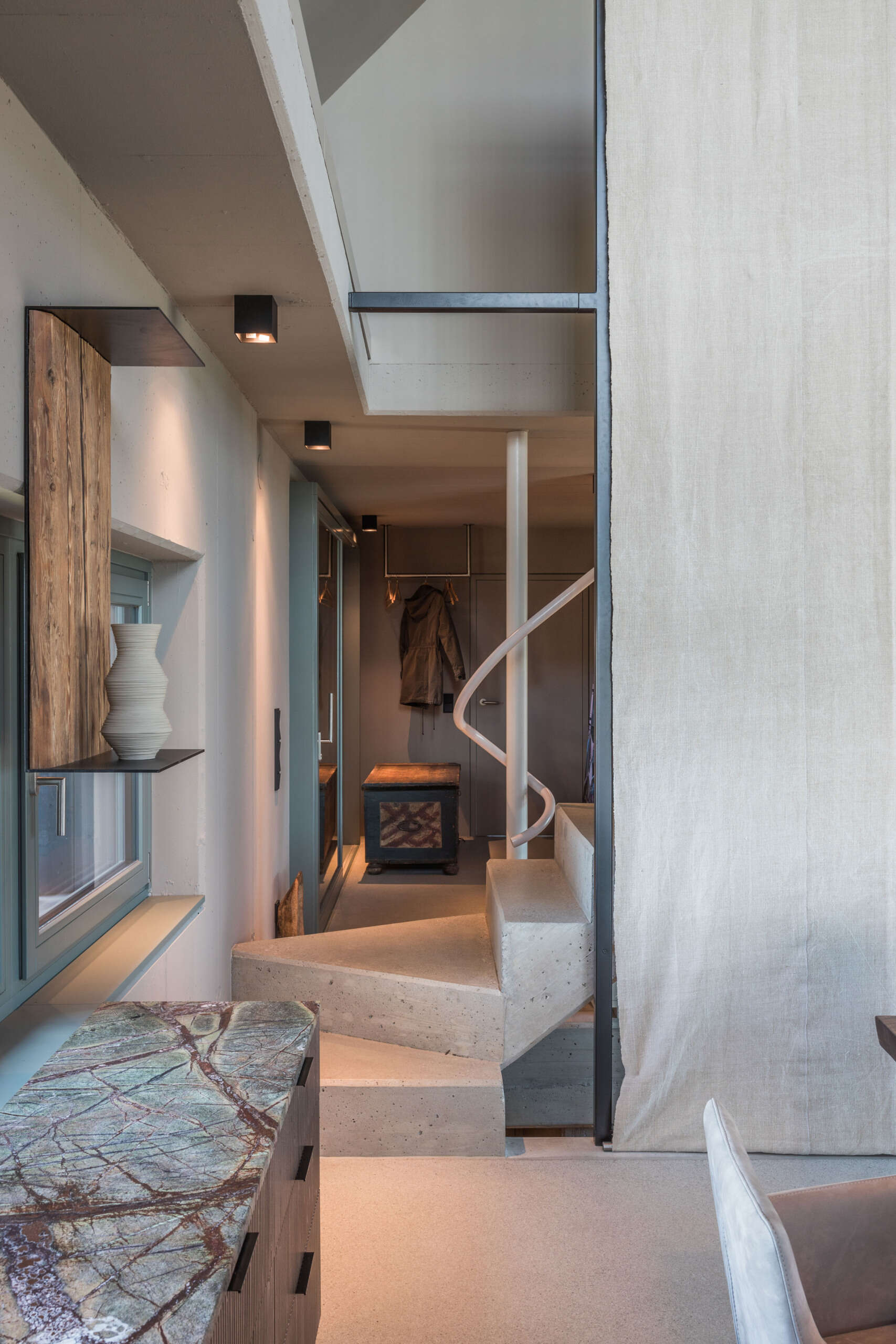 A sculptural concrete staircase in an Austrian farmhouse renovation features angular steps, a pale pink curved handrail, and a custom storage bench with upholstered seating.