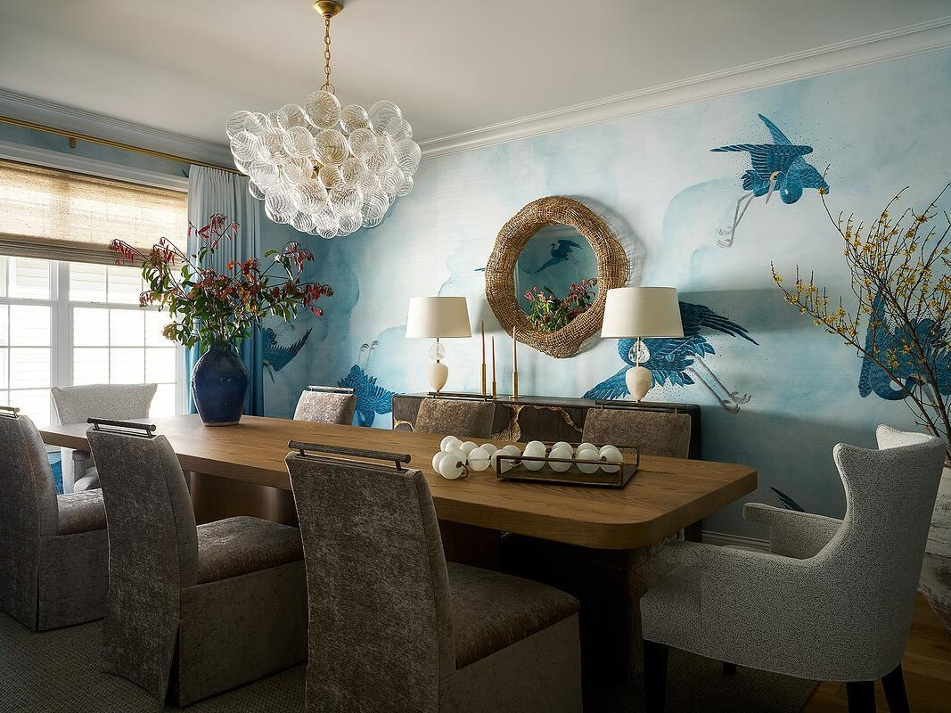 A coastal-inspired dining room featuring pale blue wallpaper with large illustrated birds, a natural wood dining table, upholstered chairs, a woven round mirror, and a sculptural glass chandelier.