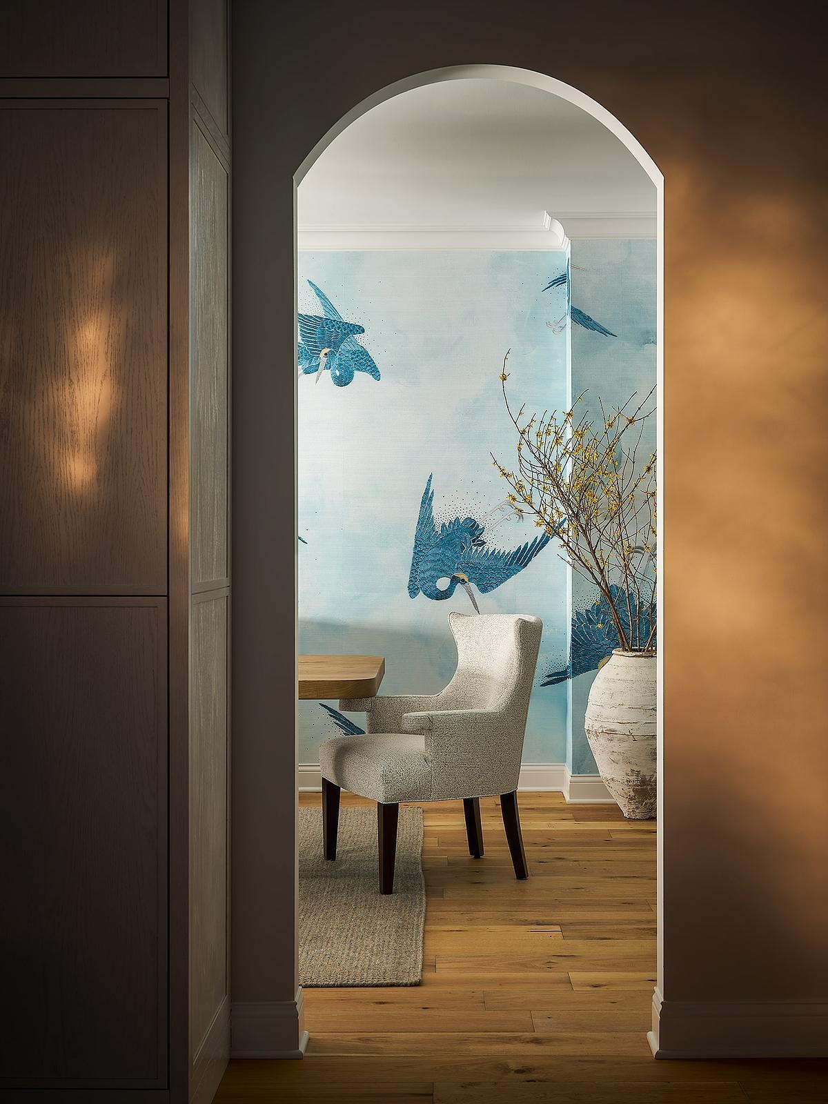 An arched doorway frames a dining room with pale blue bird-patterned wallpaper, upholstered dining chairs, natural wood flooring, and a large decorative ceramic vase.