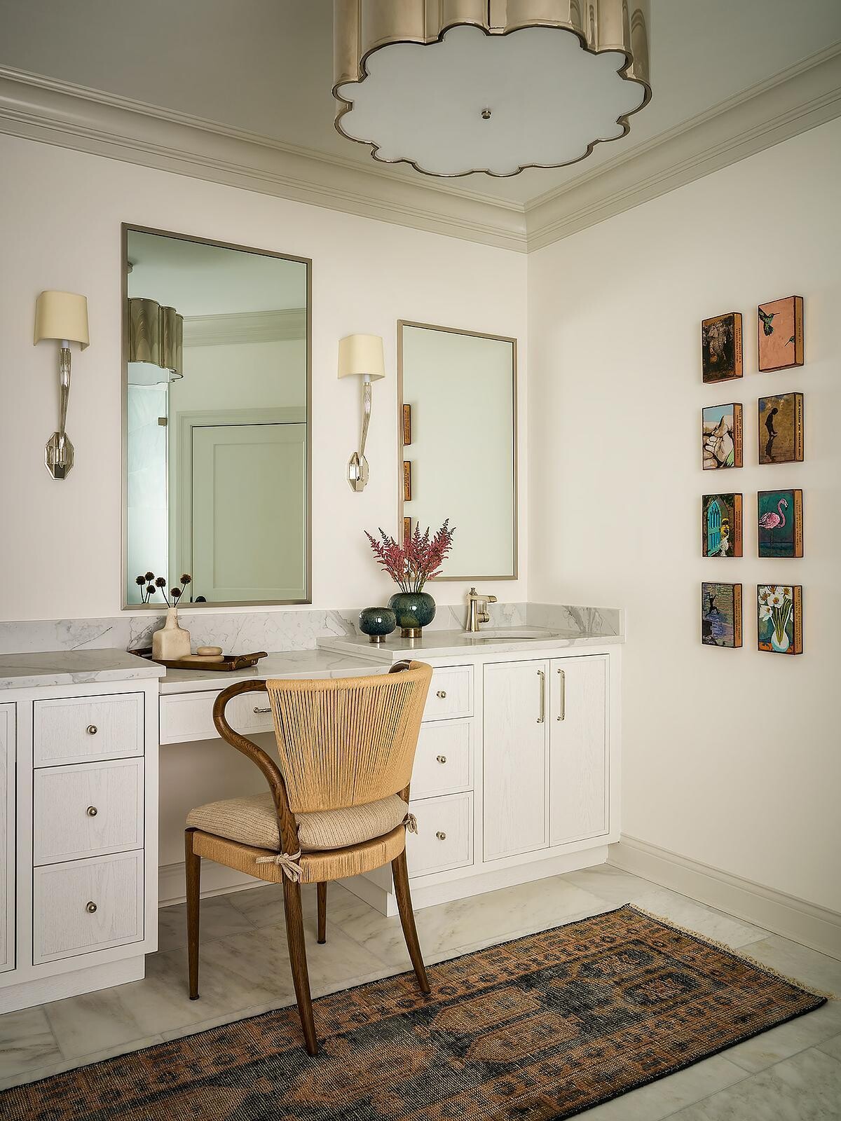A bright dressing and vanity area with white cabinetry, two tall mirrors, marble countertops, a woven accent chair, decorative wall sconces, and a gallery of small colorful framed artworks.