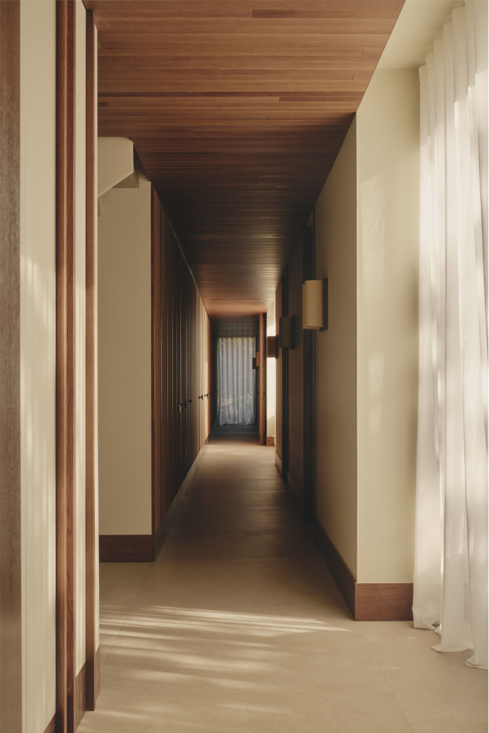 This modern hallway in Spain features natural woven wall panels, concealed doors, warm timber ceilings, sheer curtains, and courtyard windows that bring in natural light.