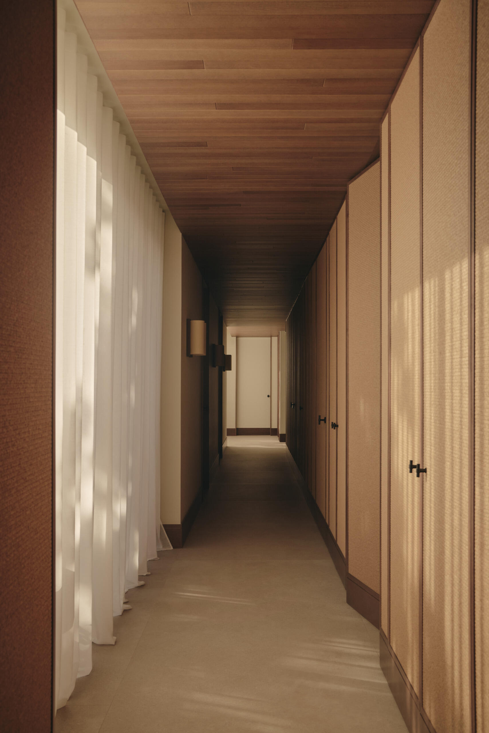 This modern hallway in Spain features natural woven wall panels, concealed doors, warm timber ceilings, sheer curtains, and courtyard windows that bring in natural light.
