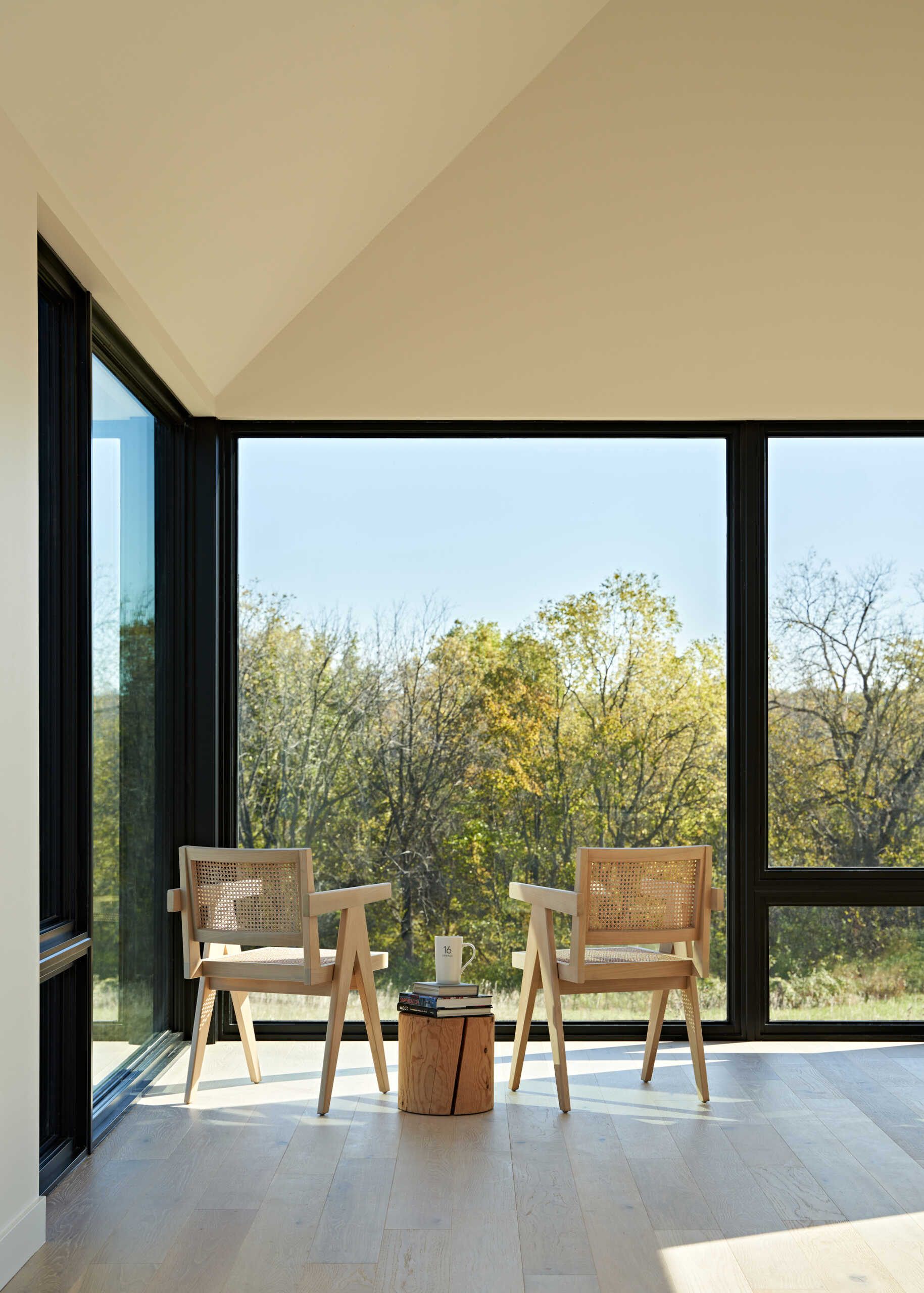 A bright modern sitting area with two light wood and woven cane chairs positioned beside floor-to-ceiling corner windows. Pale wood floors, white walls, and a small wood stump table create a simple space overlooking the trees.