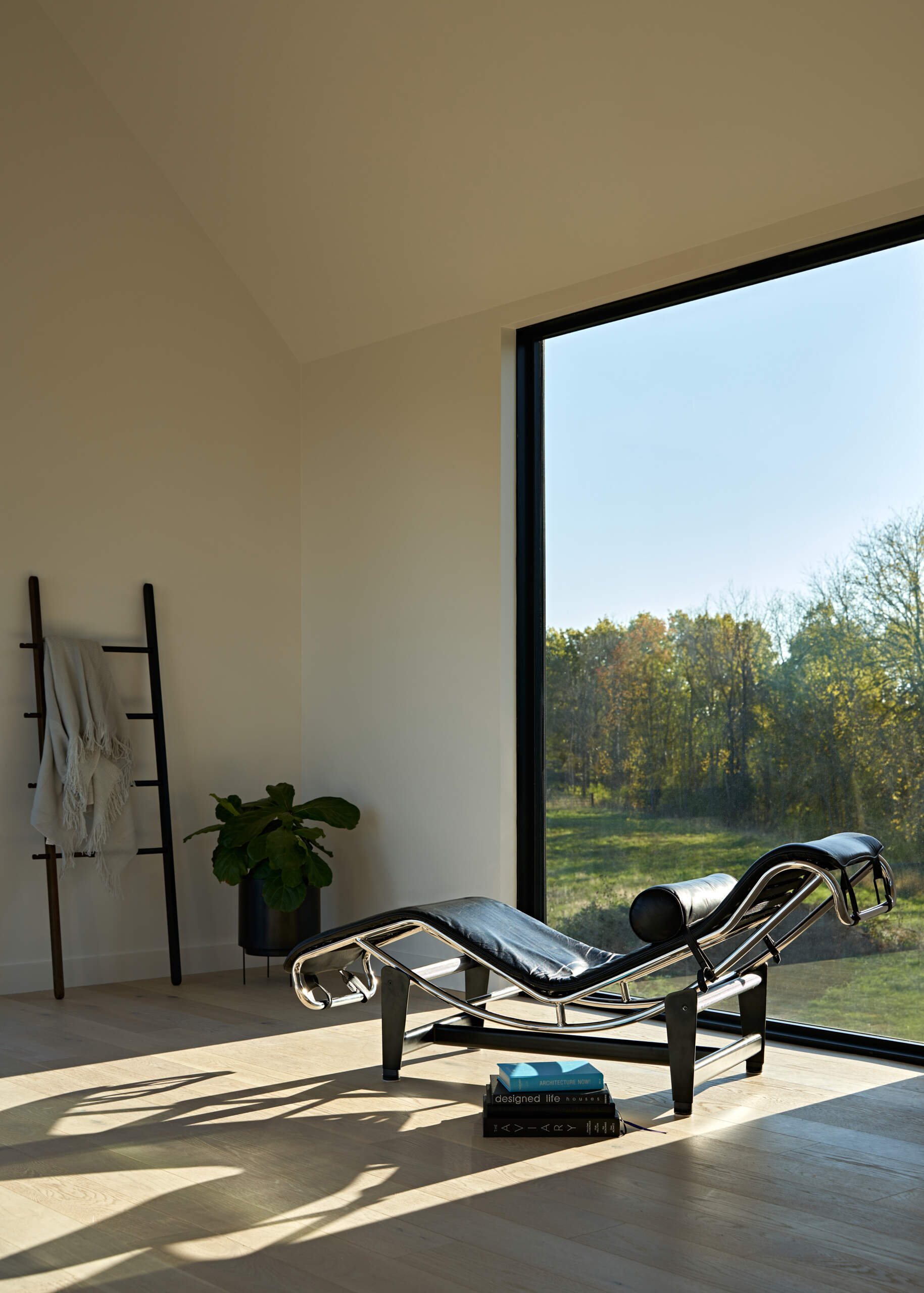 A black leather and chrome lounge chair sits beside a large floor-to-ceiling window in a minimalist modern interior. Pale wood flooring, white walls, and a decorative blanket ladder complement the view of the surrounding countryside.