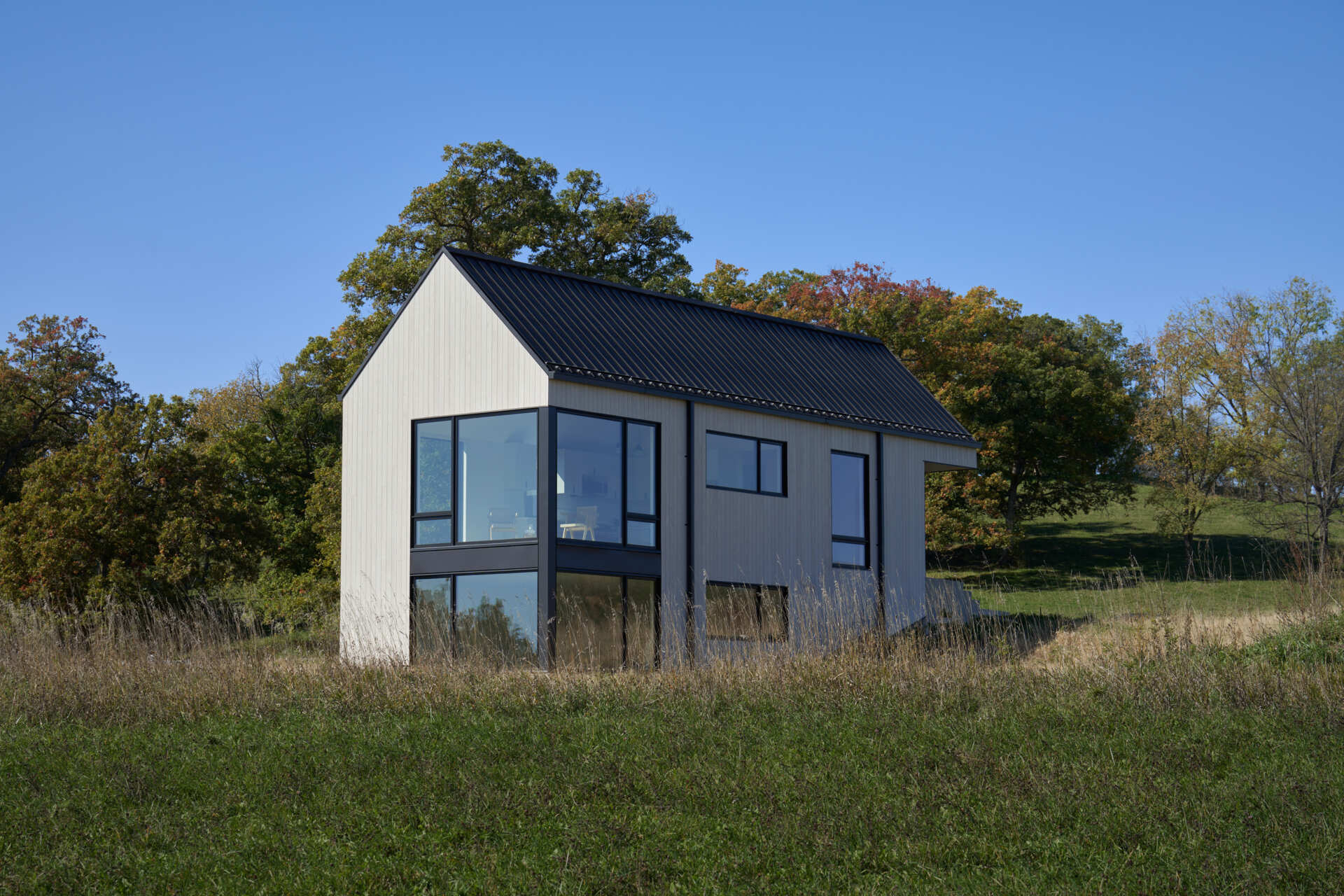 A contemporary two-story house with white vertical siding, a black metal roof, and large corner windows extending across both levels. The simple gabled architecture overlooks a grassy rural landscape surrounded by trees.