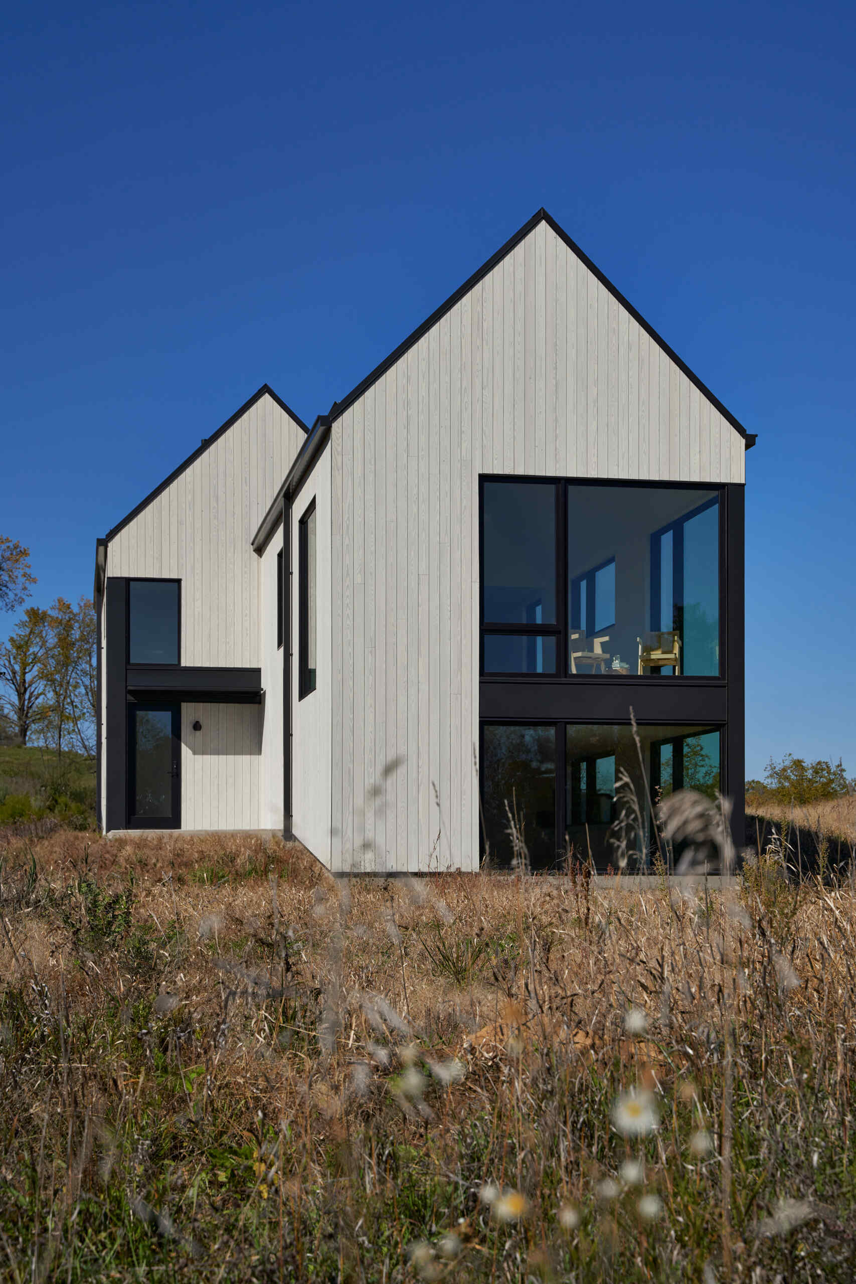 A modern pale wood cabin with steep gabled roofs and large black-framed windows arranged across two stories. Vertical wood siding and dark architectural trim contrast with the tall grass and trees surrounding the house.