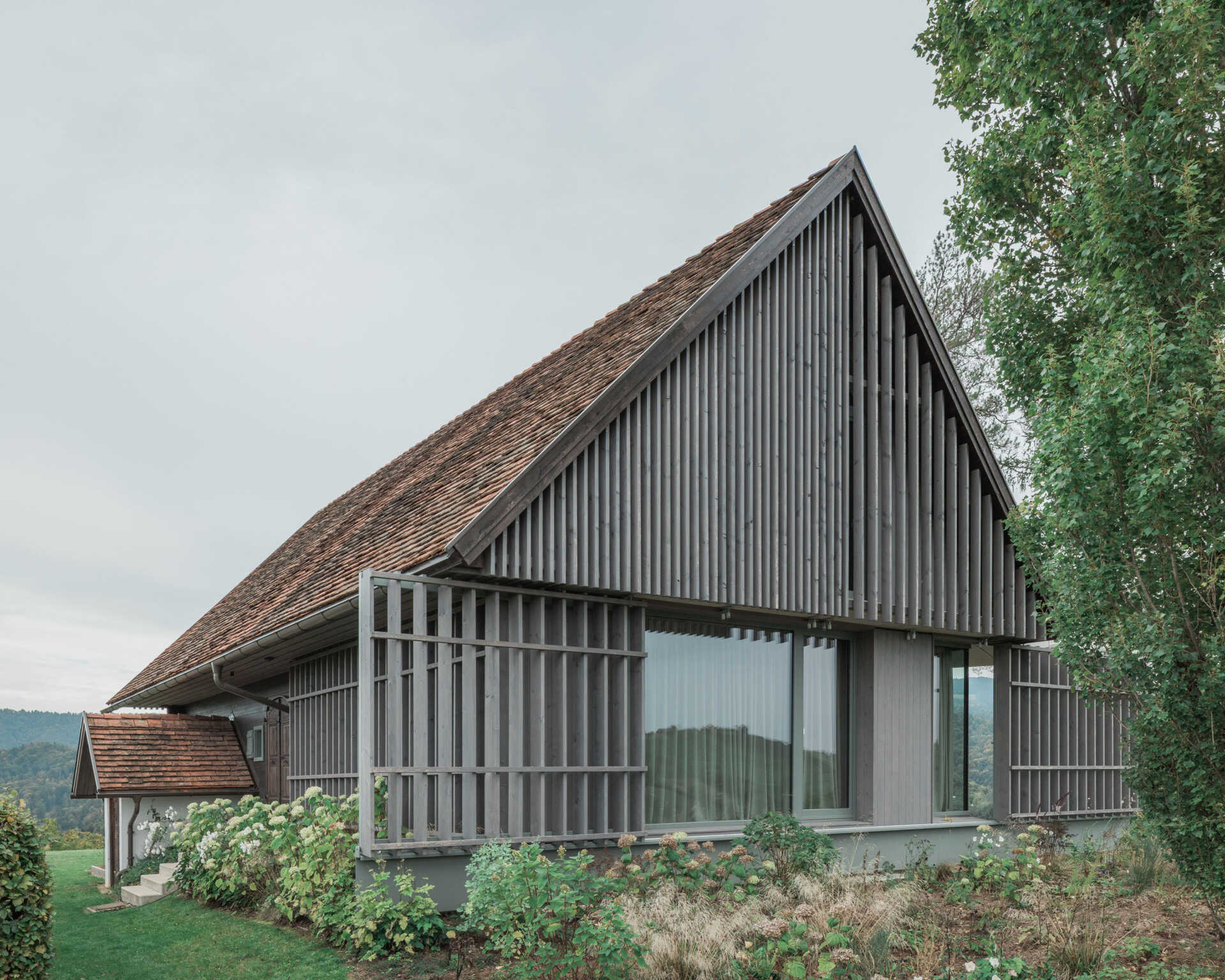 Traditional farmhouse architecture meets contemporary design in this Austrian home renovation, featuring timber screens, a pitched tile roof, modern glazing, and countryside views.