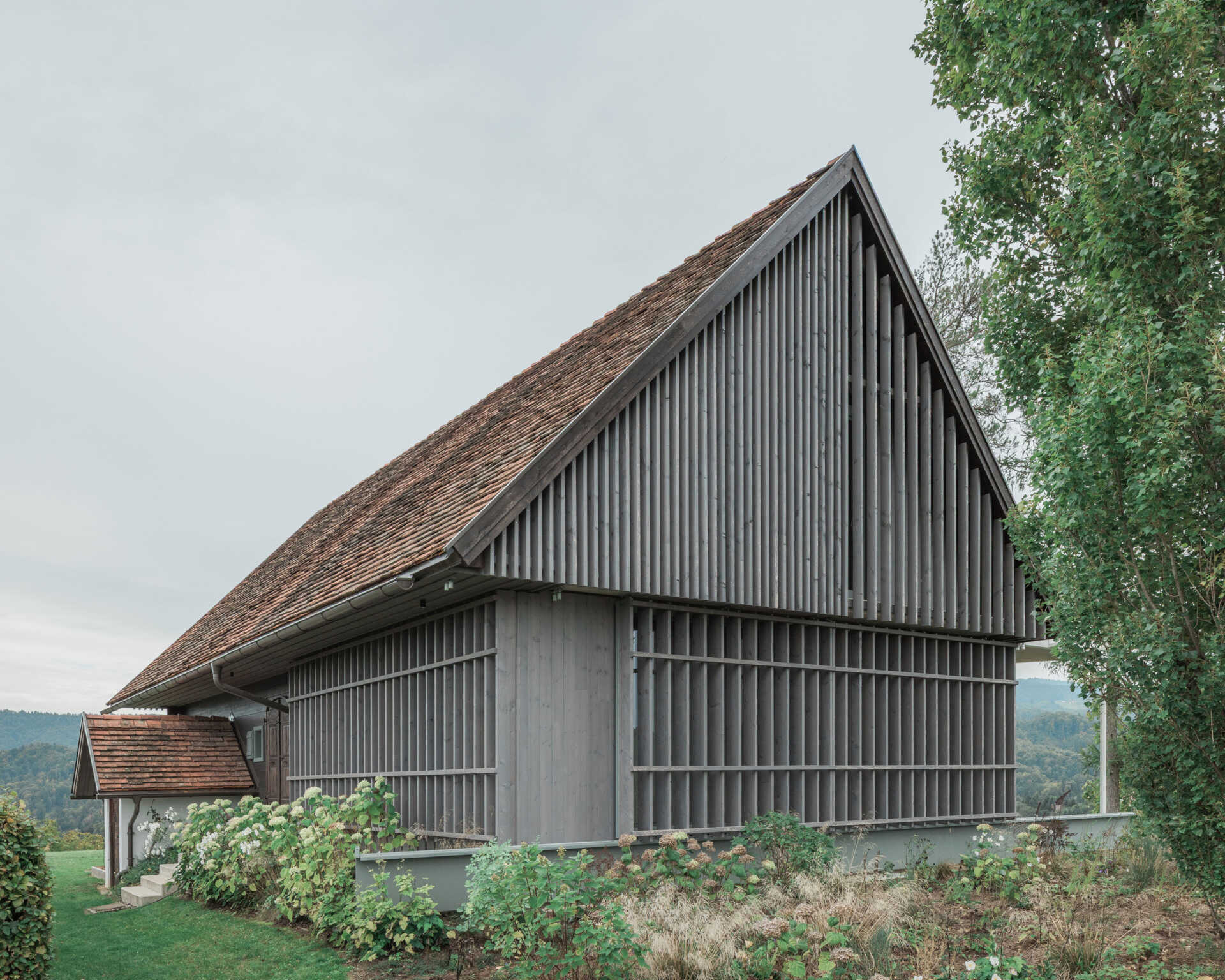 Traditional farmhouse architecture meets contemporary design in this Austrian home renovation, featuring timber screens, a pitched tile roof, modern glazing, and countryside views.