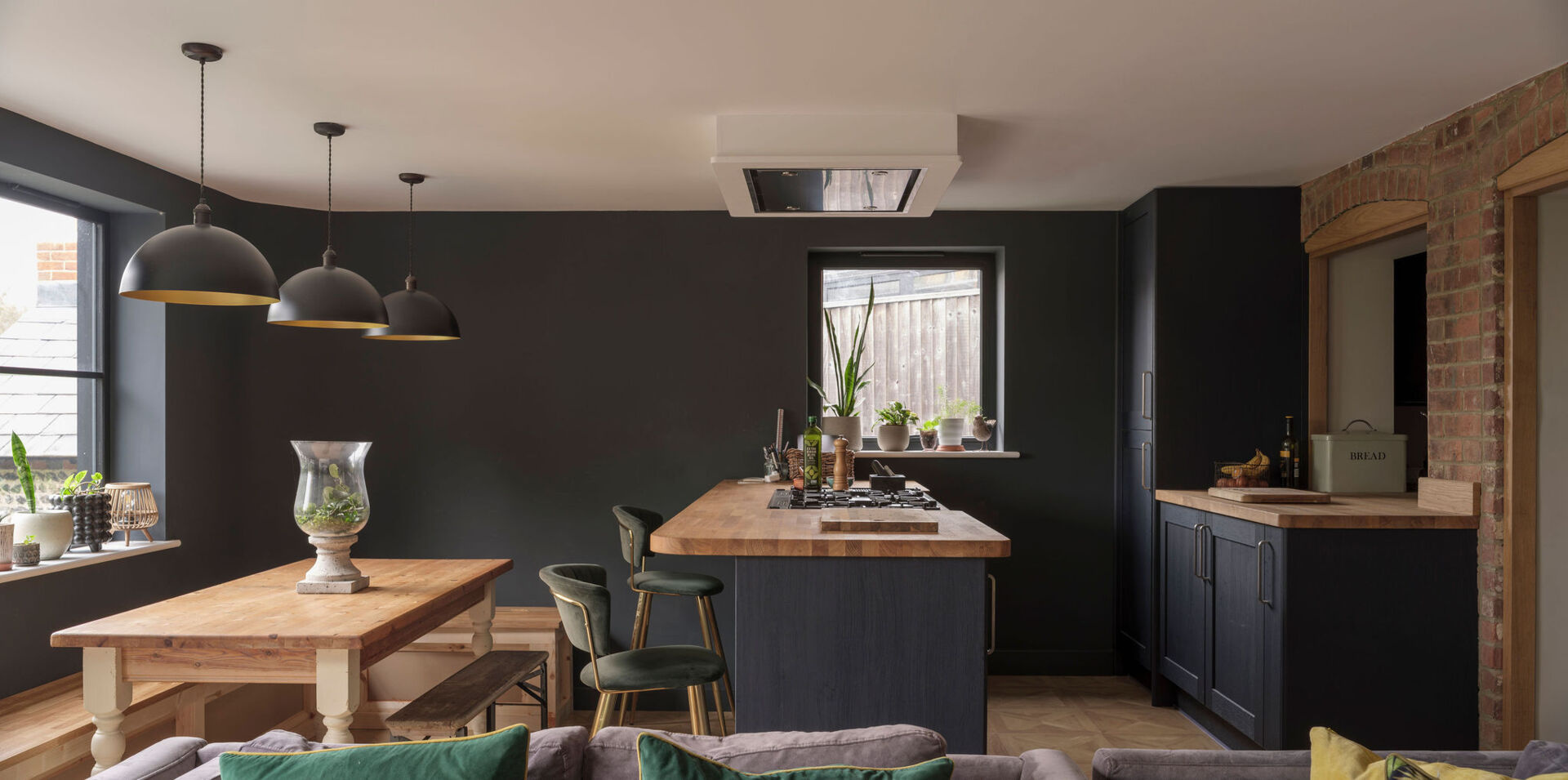 An open-plan kitchen and dining area pairs dark cabinets, natural wood countertops, a kitchen island, and rustic exposed brick arches in a renovated English cottage.