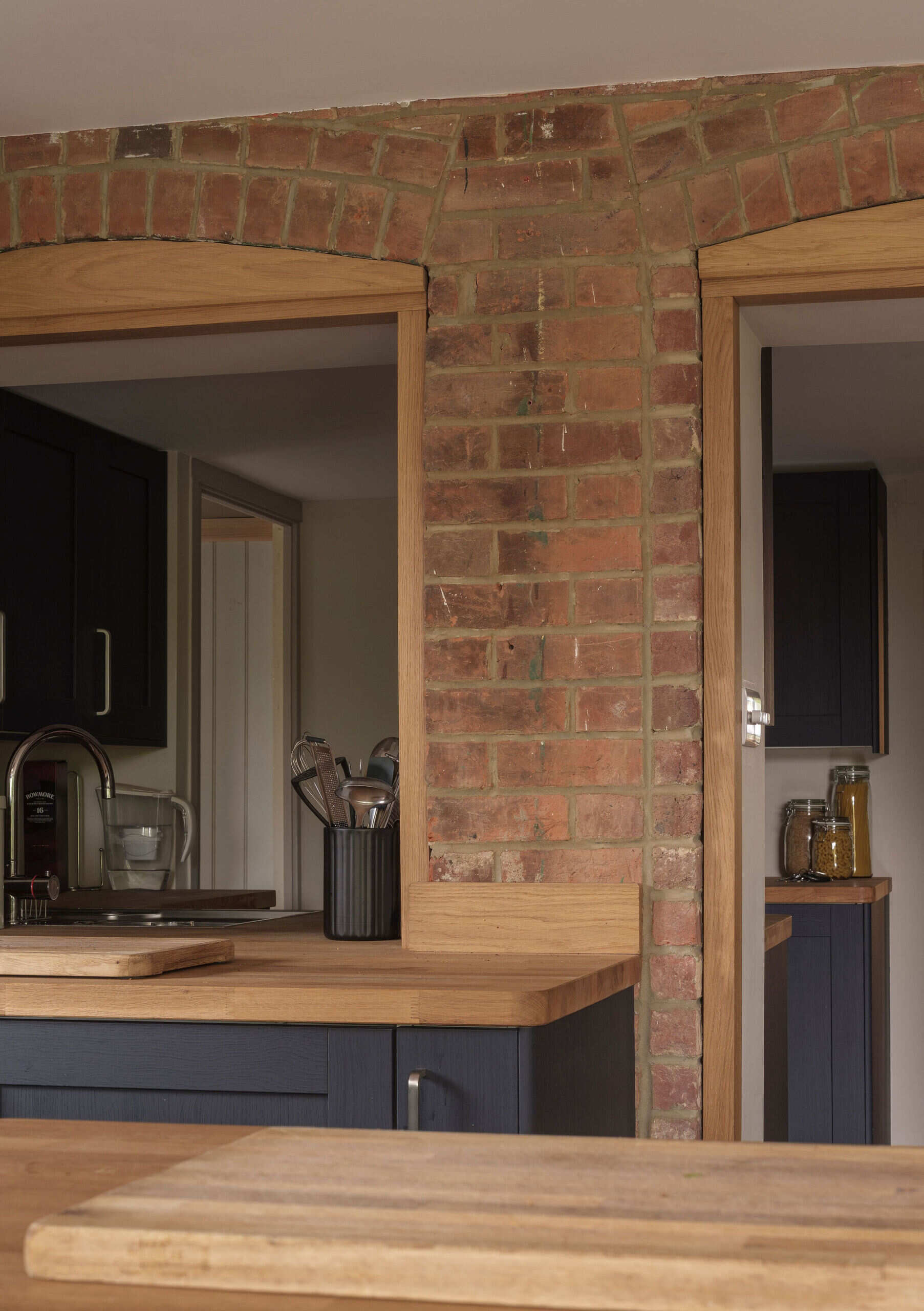 An open-plan kitchen and dining area pairs dark cabinets, natural wood countertops, a kitchen island, and rustic exposed brick arches in a renovated English cottage.