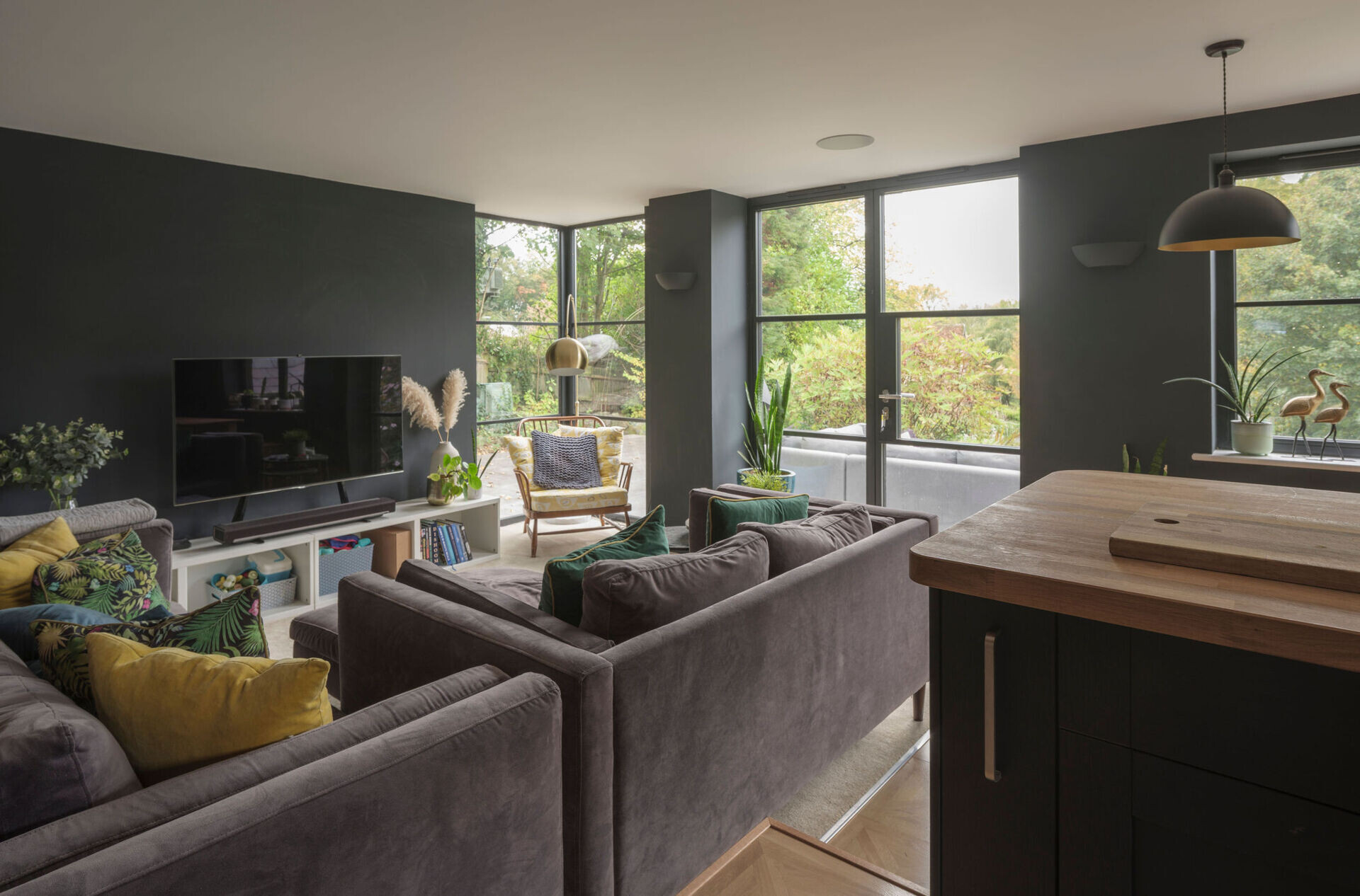A cozy contemporary living room with charcoal walls, gray sofas, colorful cushions, and large garden-facing windows forms part of a modern cottage extension.