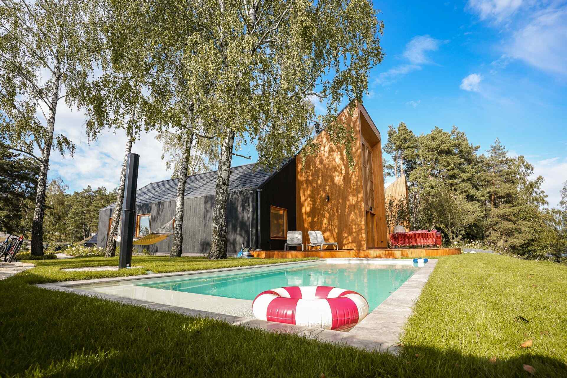 Discover the exterior of Kalbornia Lake & Pool House in Poland, featuring an angular timber-clad house, heated outdoor swimming pool, wooden deck, and landscaped grounds surrounded by trees.