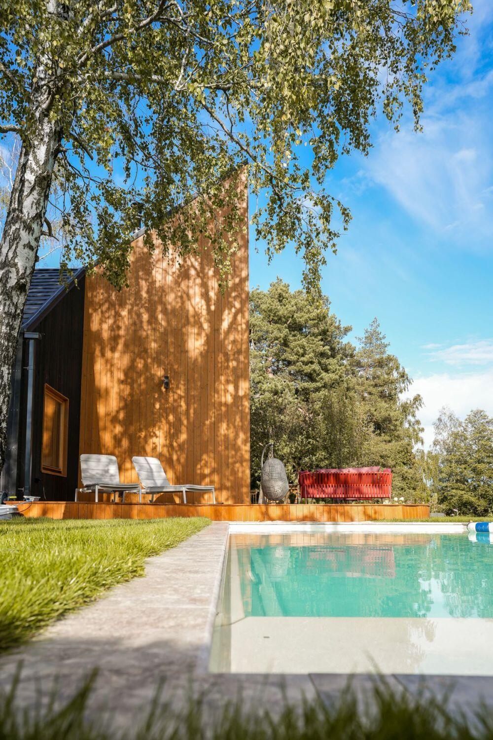 Discover the exterior of Kalbornia Lake & Pool House in Poland, featuring an angular timber-clad house, heated outdoor swimming pool, wooden deck, and landscaped grounds surrounded by trees.