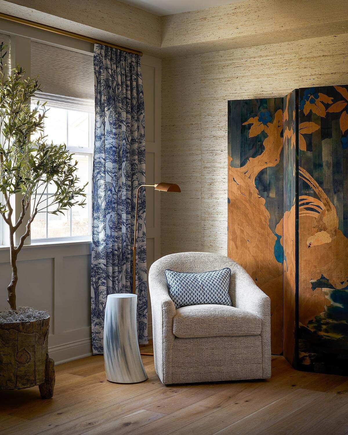 A cozy bedroom reading corner featuring an antique painted folding screen, a curved upholstered armchair, blue and white patterned curtains, a brass floor lamp, and textured neutral wallpaper.