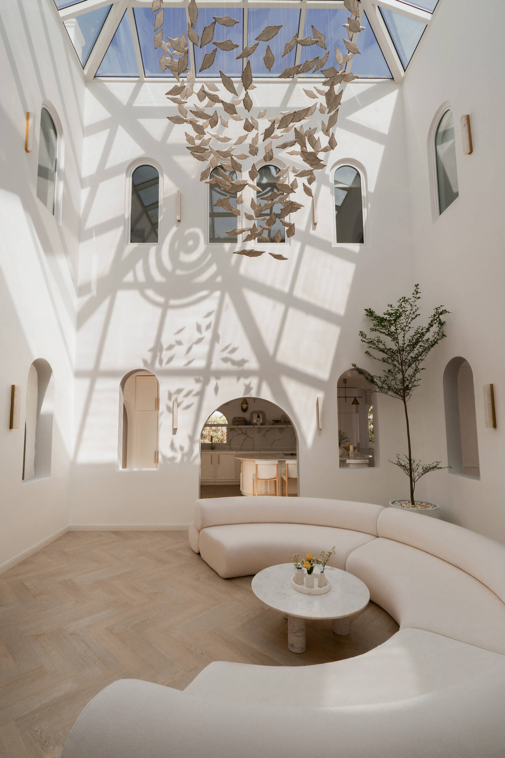 Discover the central courtyard inside The White Arcade, a Dubai villa renovated by D’Ora Tokai Designs. Featuring double-height ceilings, a glass roof, sculptural arches, a bespoke C-shaped sofa, indoor trees, and a floating leaf installation, this unique living space is filled with natural light.