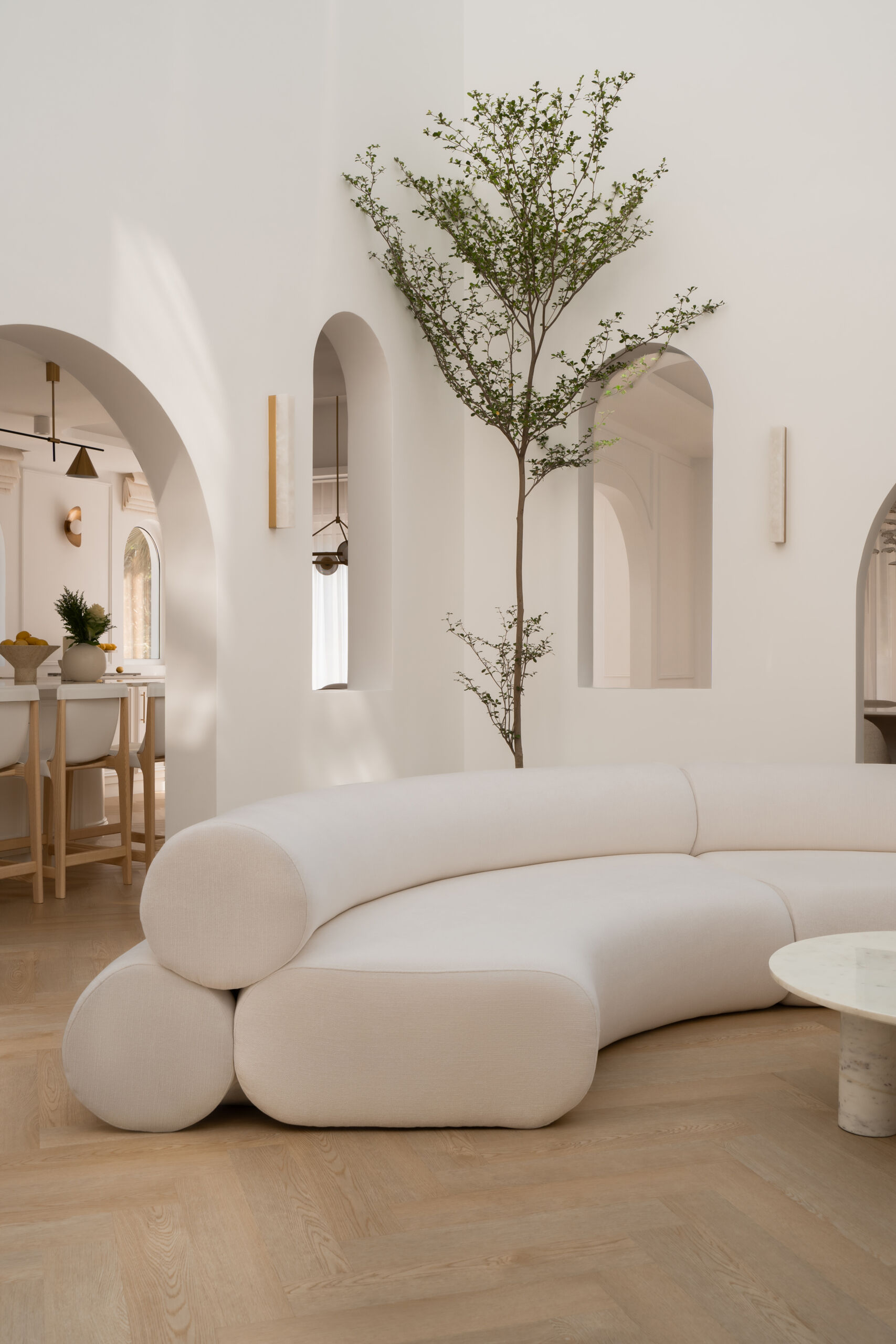Discover the central courtyard inside The White Arcade, a Dubai villa renovated by D’Ora Tokai Designs. Featuring double-height ceilings, a glass roof, sculptural arches, a bespoke C-shaped sofa, indoor trees, and a floating leaf installation, this unique living space is filled with natural light.